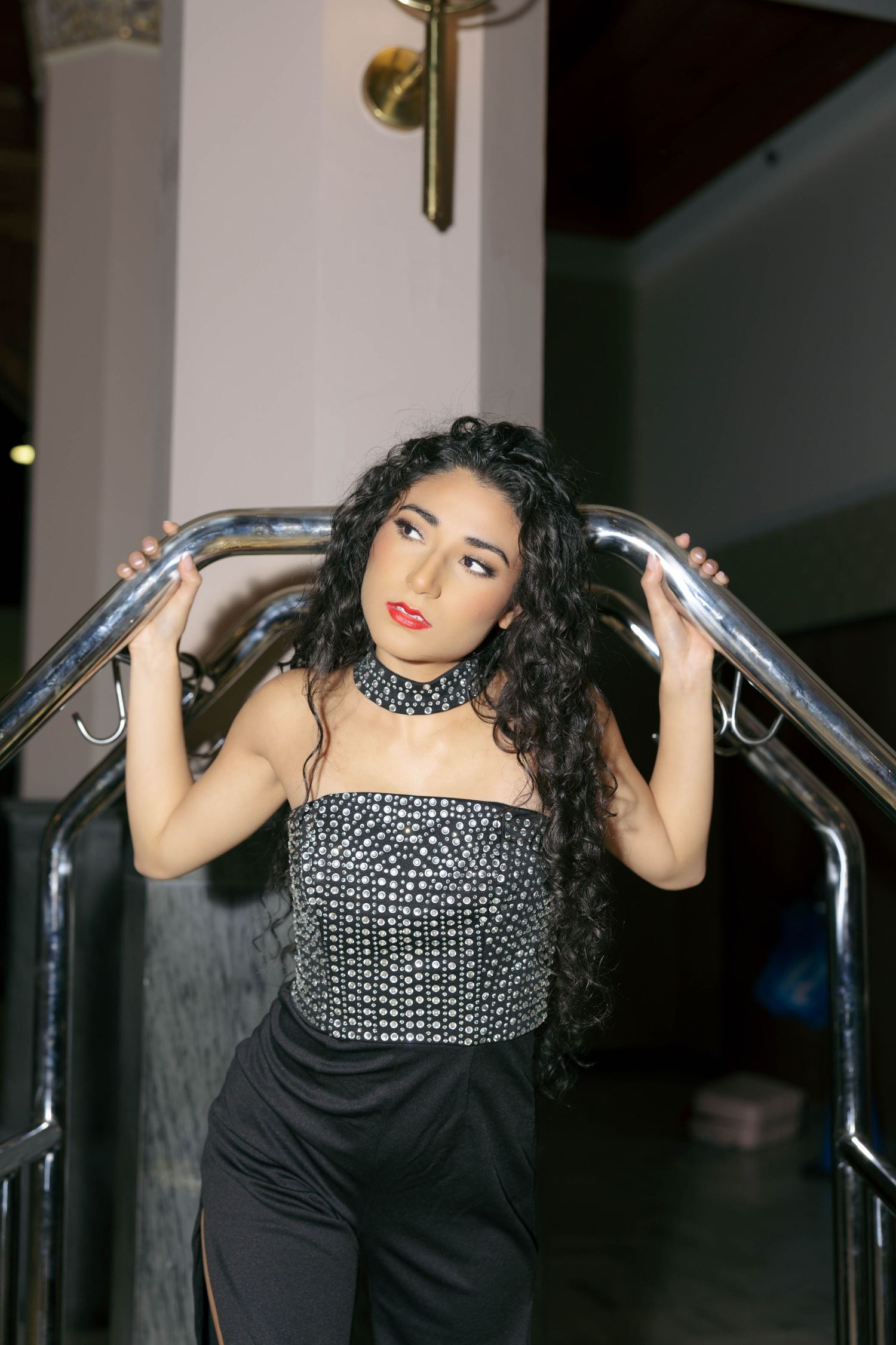 Young woman with curly hair in studded strapless top and black pants posing on staircase railing.