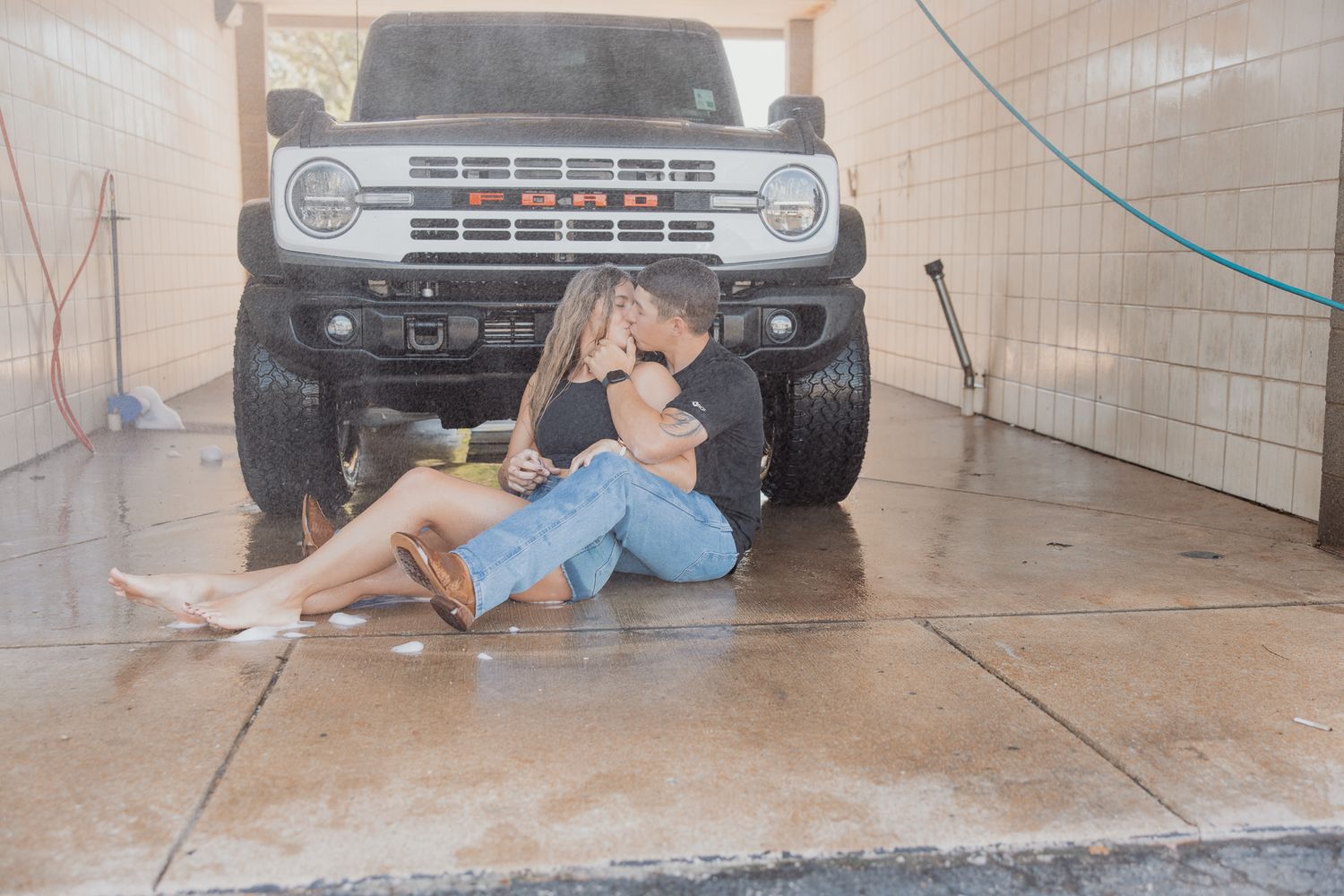 Casual couple lounging on garage floor with vintage Ford Bronco in background.