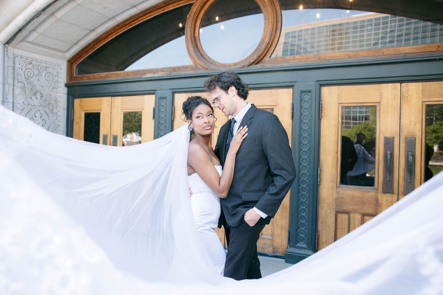 Romantic couple poses before elegant venue entrance as bride's long white veil flows beautifully in breeze.