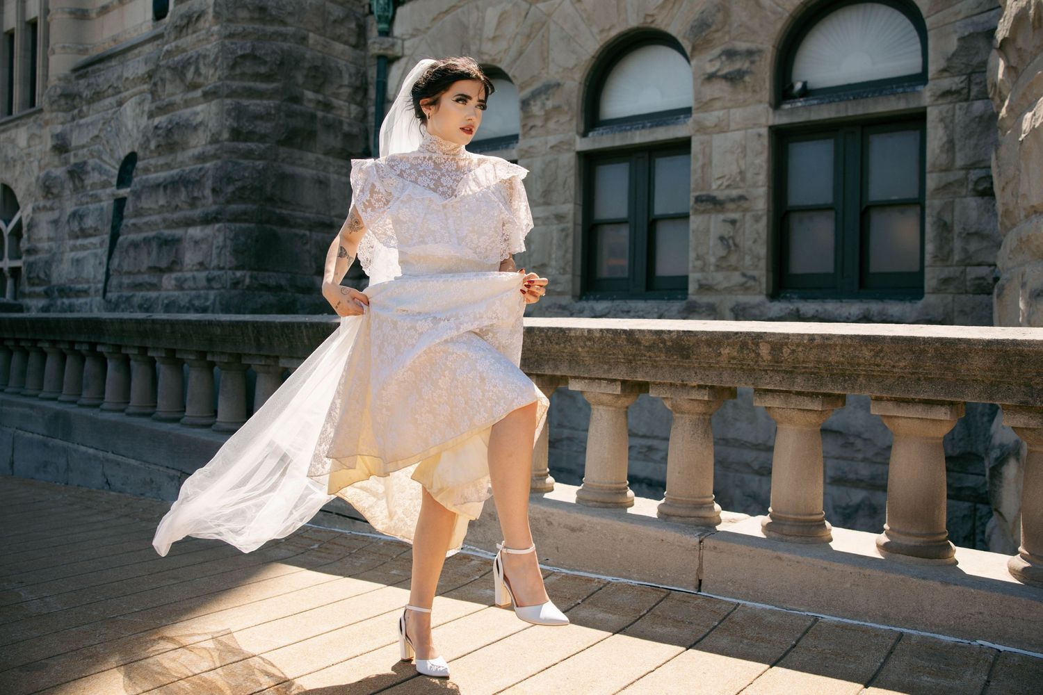 Bride in white lace dress with veil flowing in wind poses on sunlit stone balcony of historic building.