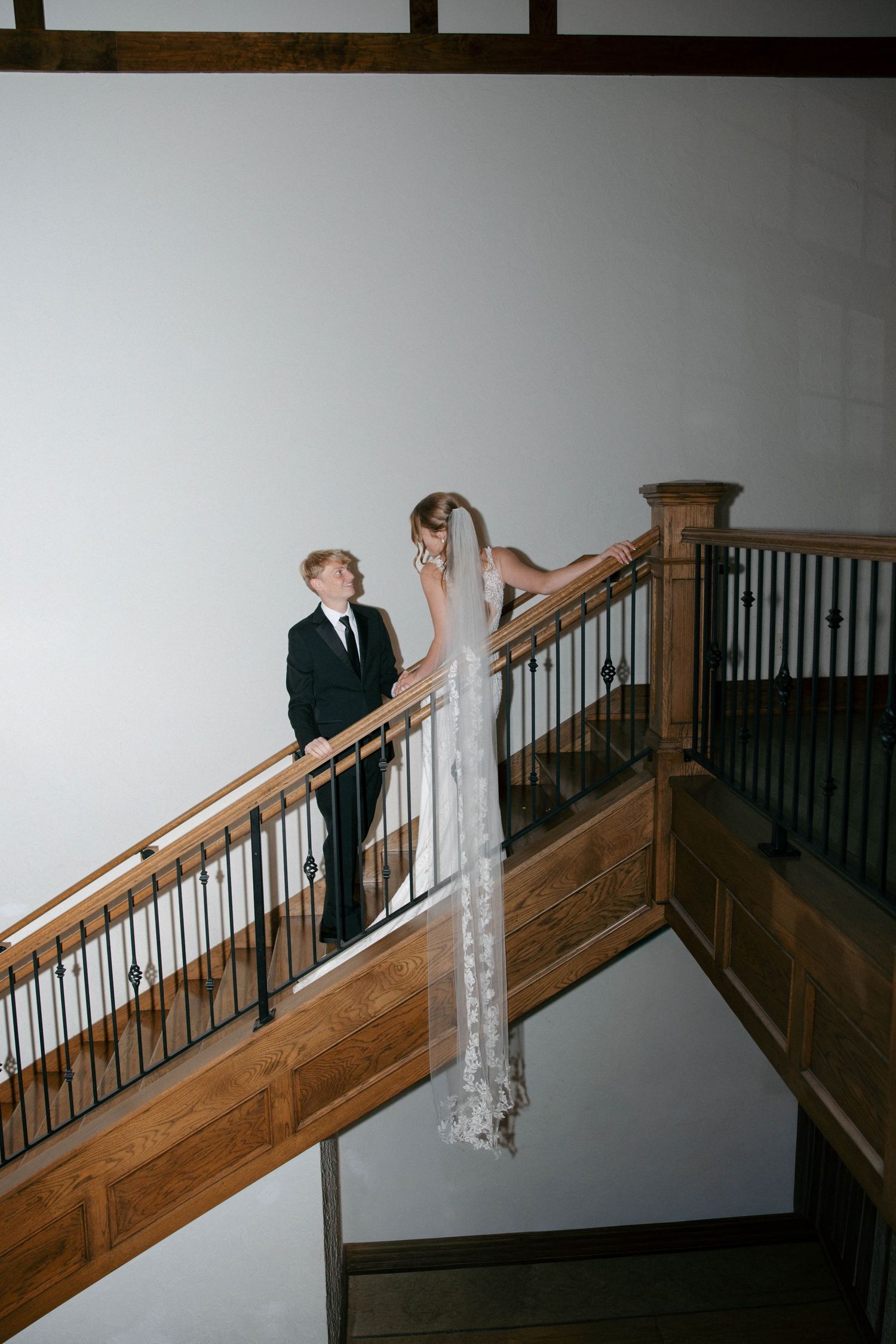 Bride in lace gown with veil and groom in black suit share a moment on elegant wooden staircase.