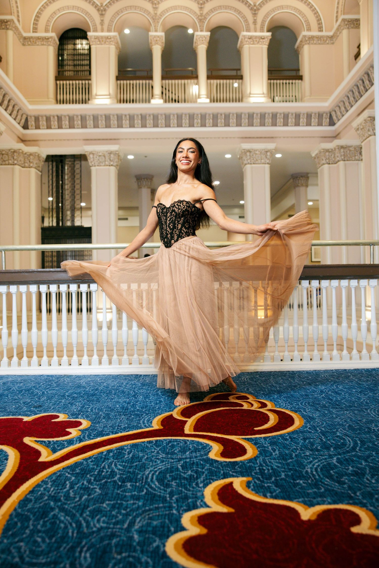 Woman in black lace corset and flowing beige tulle skirt twirling in elegant grand hall with columns.