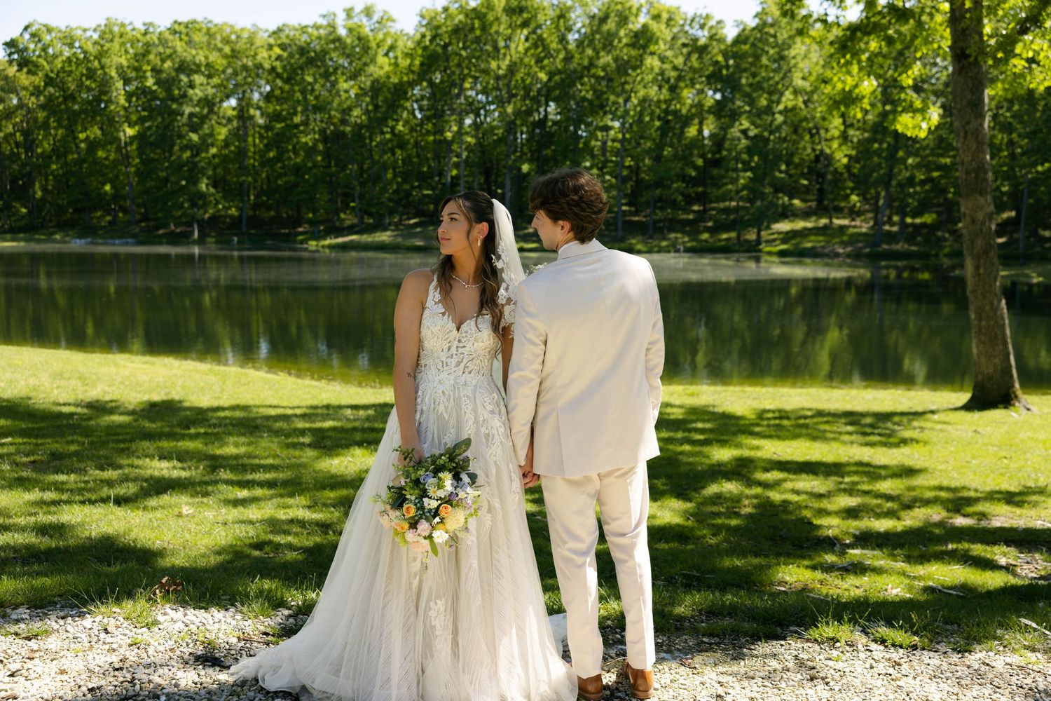 Bride and groom holding hands by a serene pond surrounded by lush green trees on their wedding day.