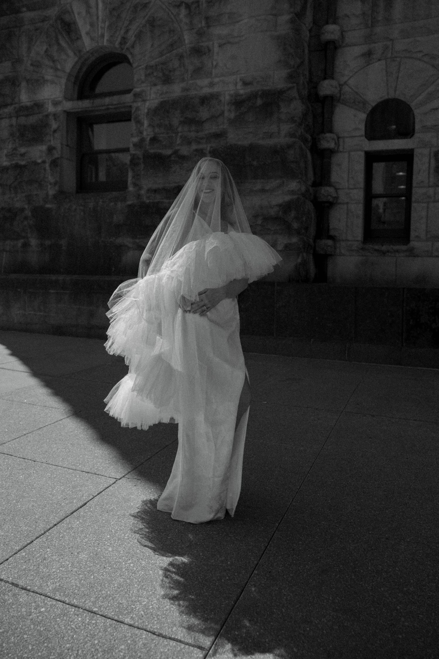 Black and white artistic photo of bride in white gown with flowing tulle veil outside historic building.