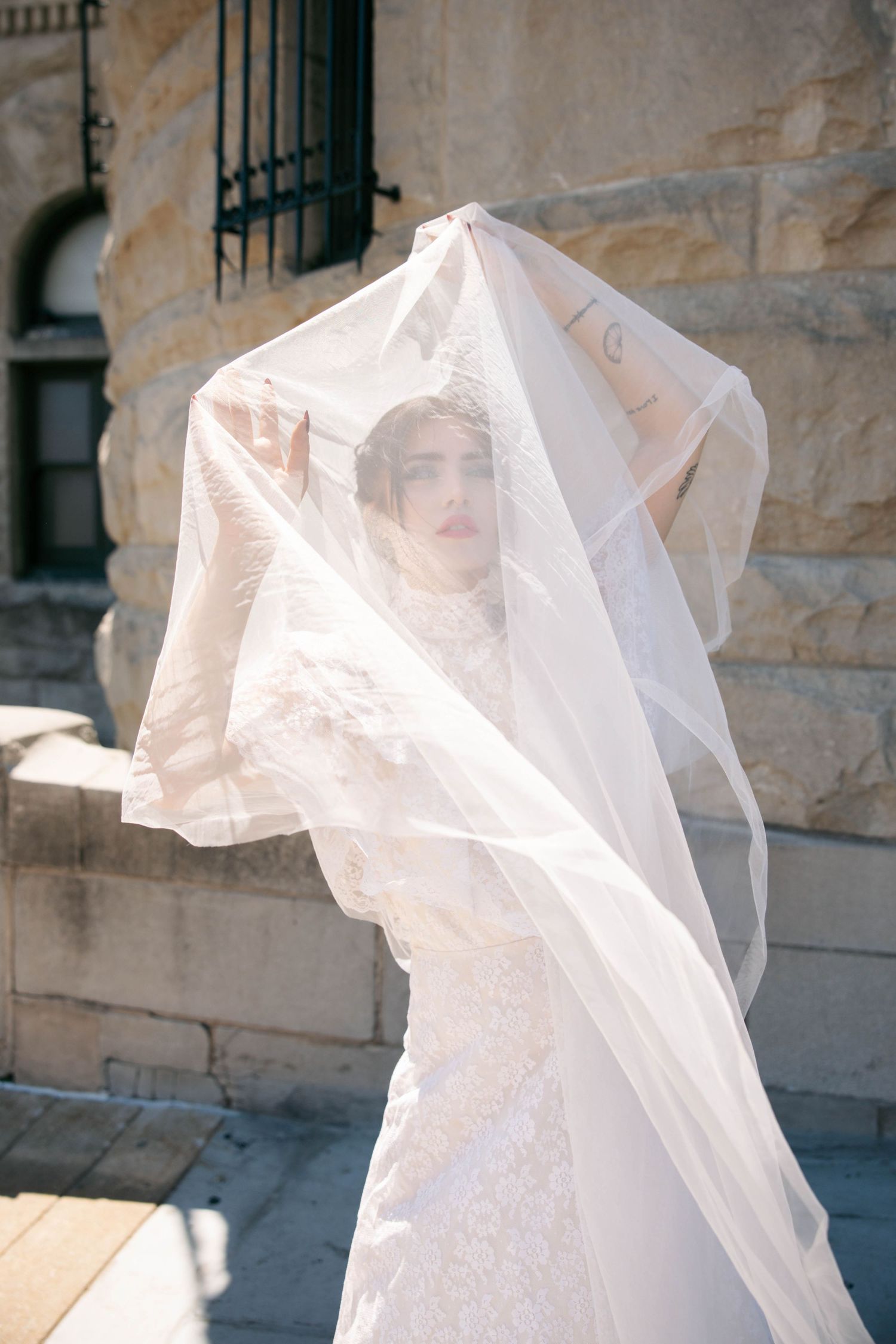 Bride in lace wedding gown holds flowing veil overhead outside stone building in sunlight.