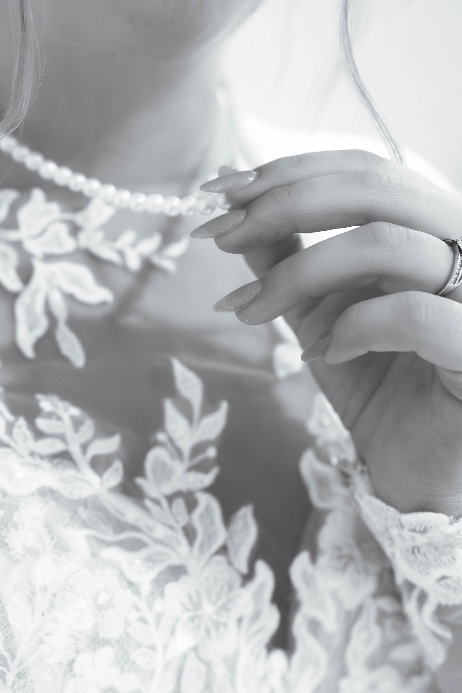 Close-up of a bride in lace wedding gown touching pearl necklace, wearing diamond ring, black and white photo.