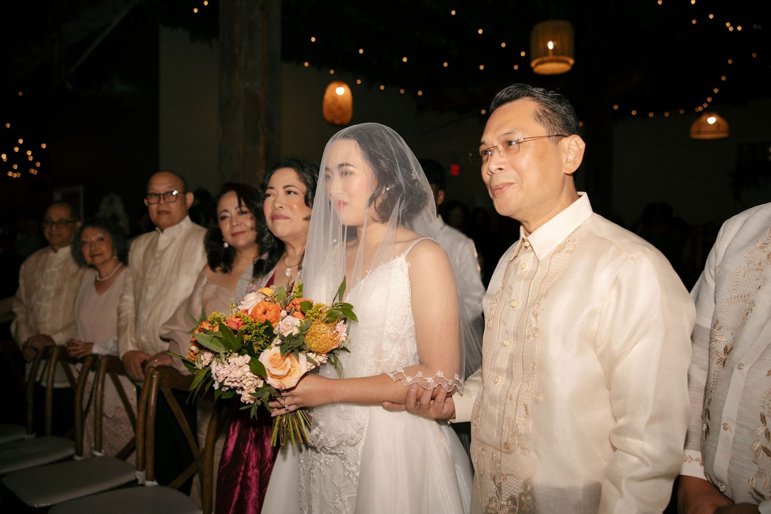 Wedding guests in traditional white attire stand together during an evening ceremony.