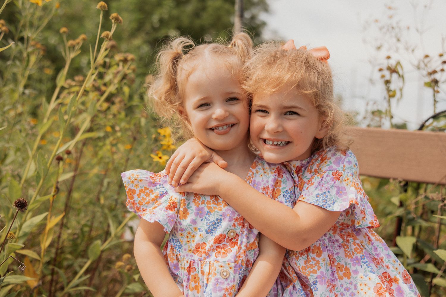 Two people embrace while wearing floral and brown outfits in a natural outdoor setting.