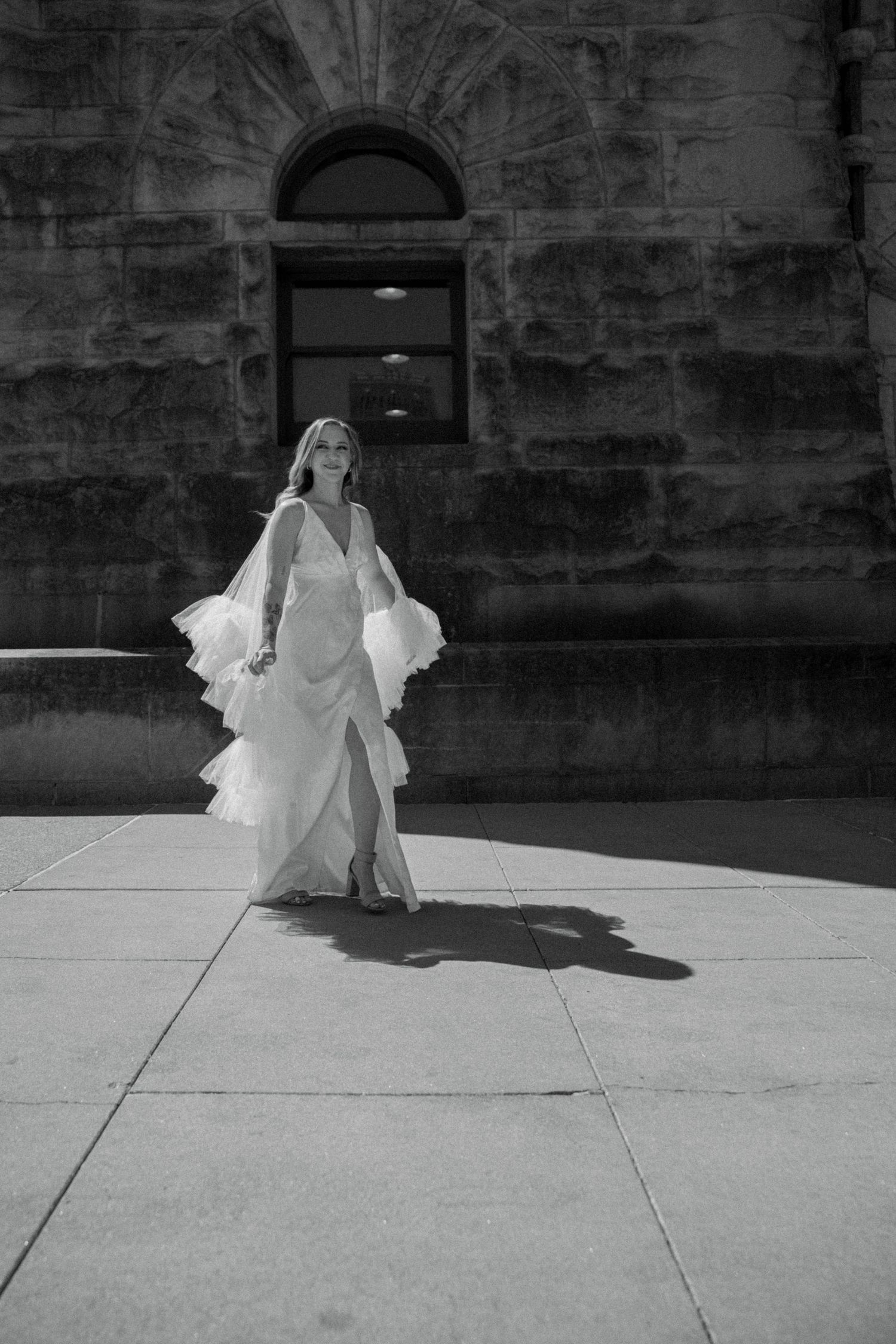 Bride in elegant white gown with ruffled cape walking on sunlit sidewalk beside historic stone architecture.