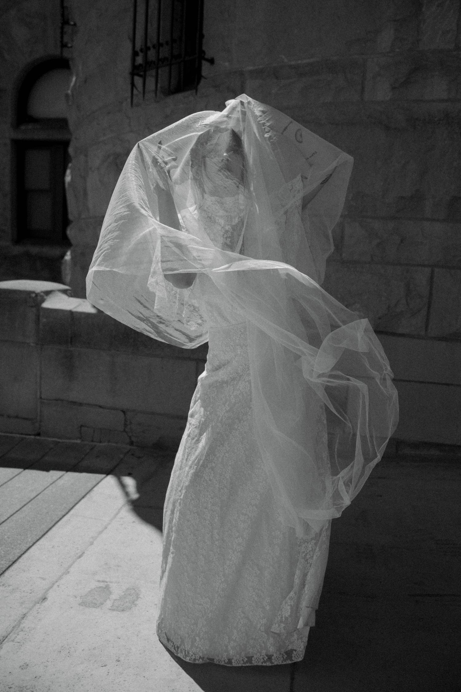 Black and white photo of a bride in a lace wedding dress with veil blowing over her face outdoors.