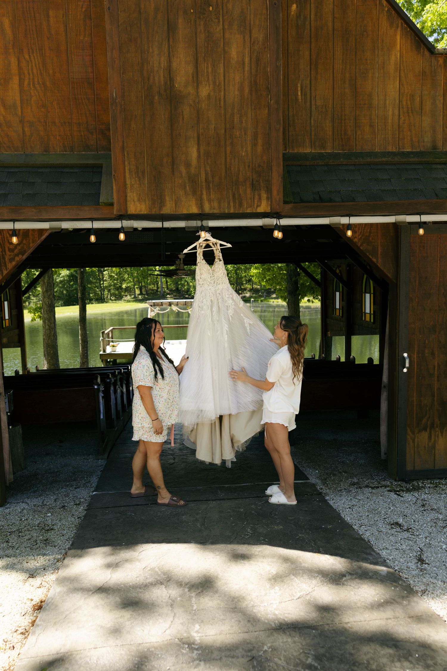 Two women in white outfits admire a wedding dress hanging in a rustic wooden barn with a lake view.
