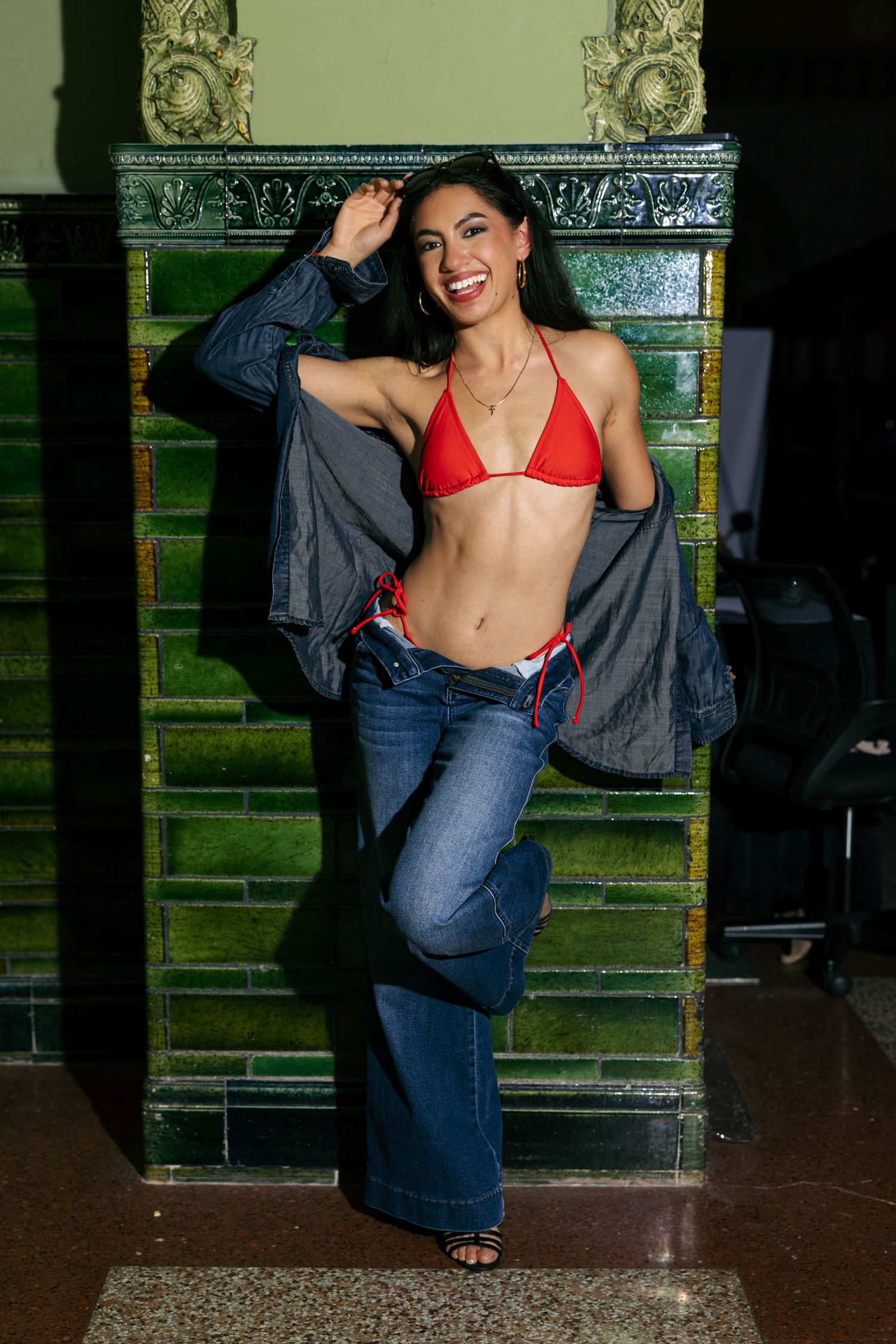 Smiling woman in red bikini top and flared jeans posing by ornate green tiled wall.