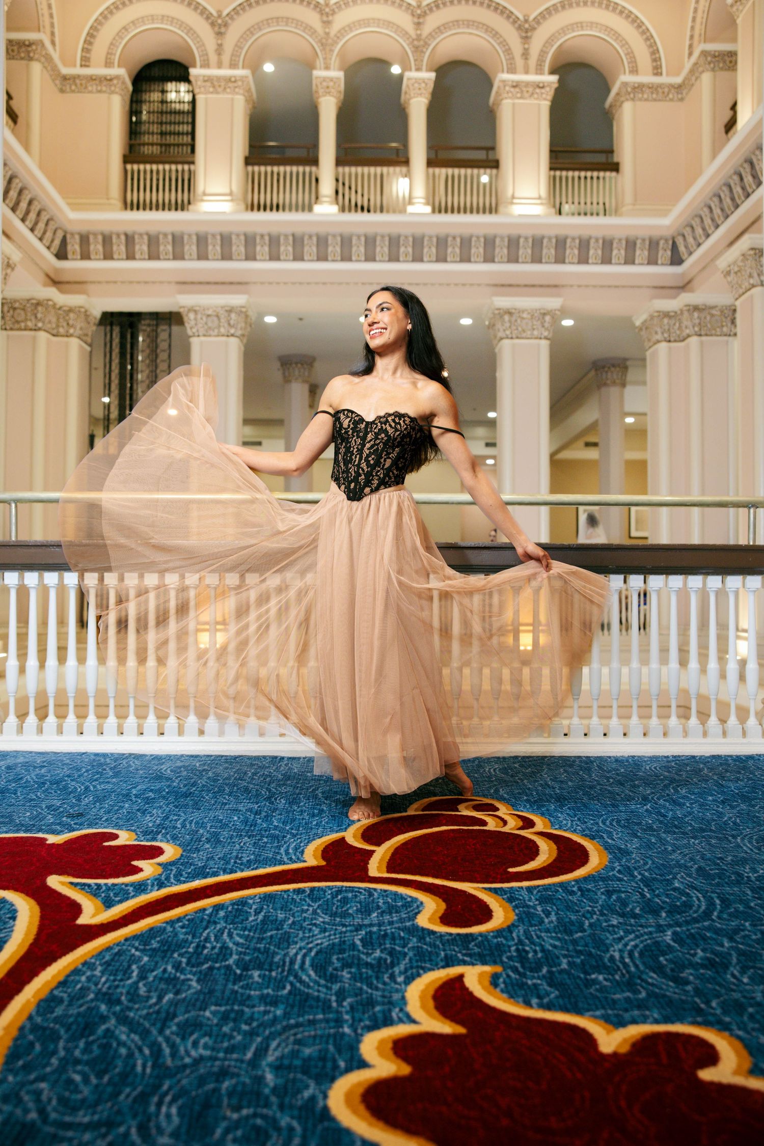 Elegant woman in black lace and nude tulle gown twirling in a grand ornate ballroom with blue carpet.