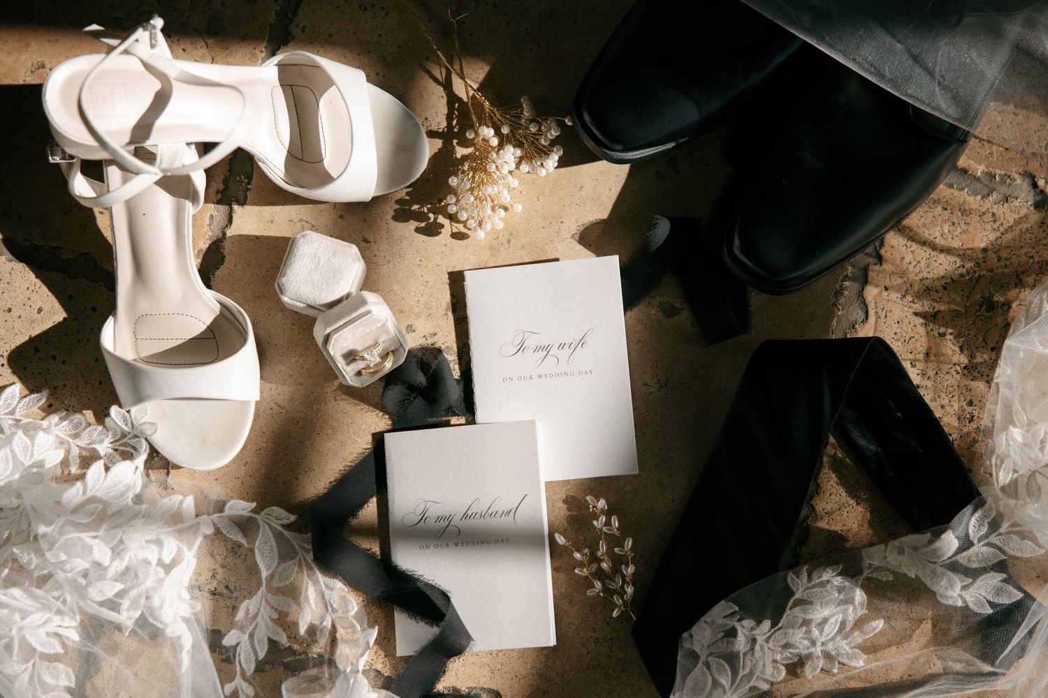 Flat lay of white bridal heels, wedding cards, groom accessories, and floral details on a textured floor.