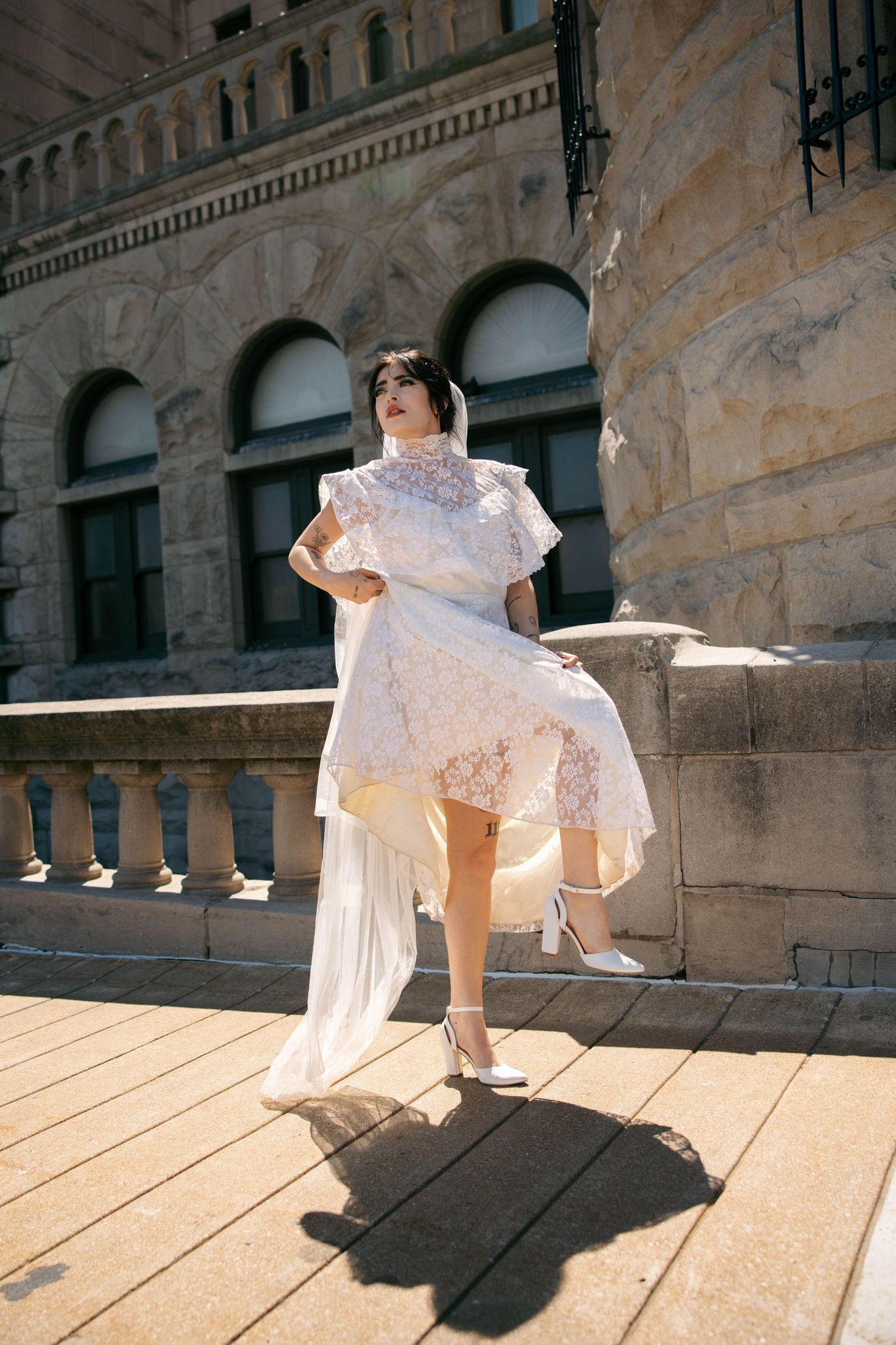 Woman in white lace dress poses on stone balcony of historic building in sunlight.