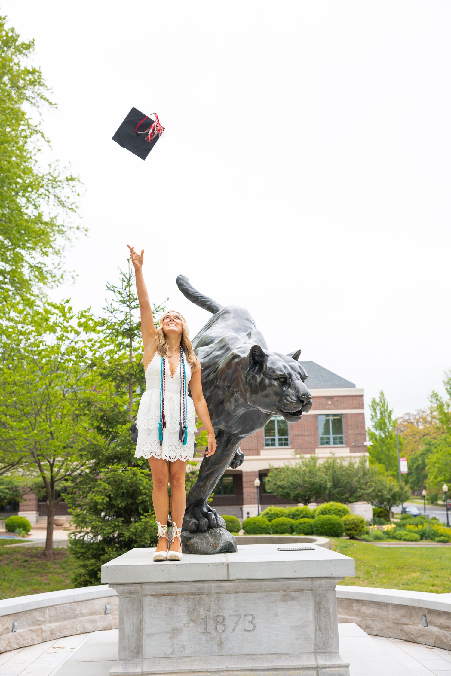 Graduate tossing cap in the air while standing on campus statue.