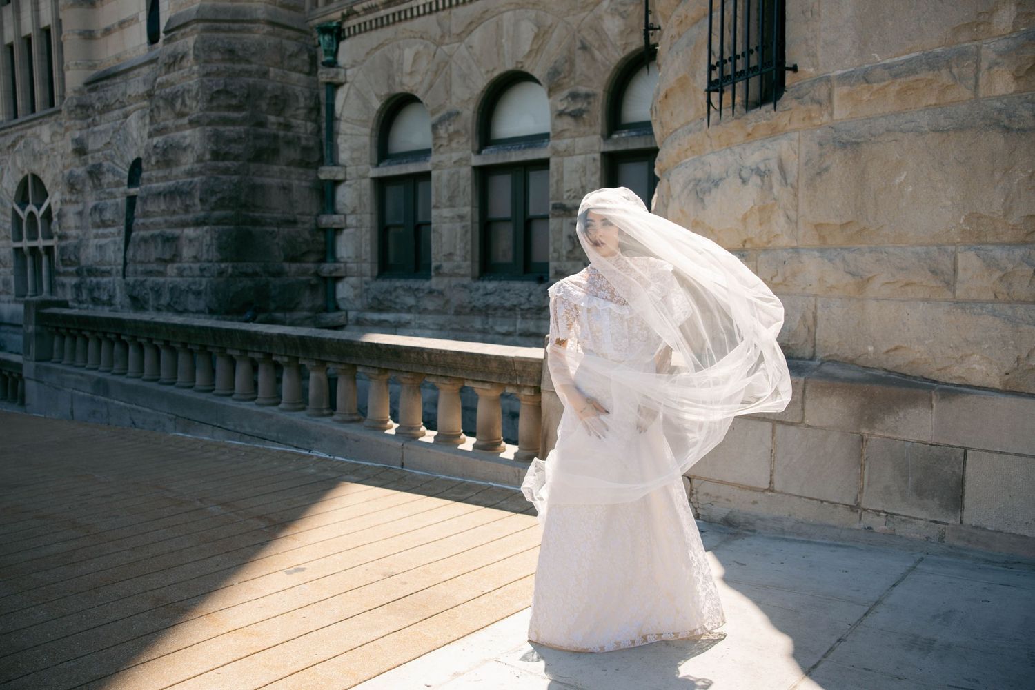 Bride in white wedding dress and flowing veil standing on sunny terrace of historic stone building.