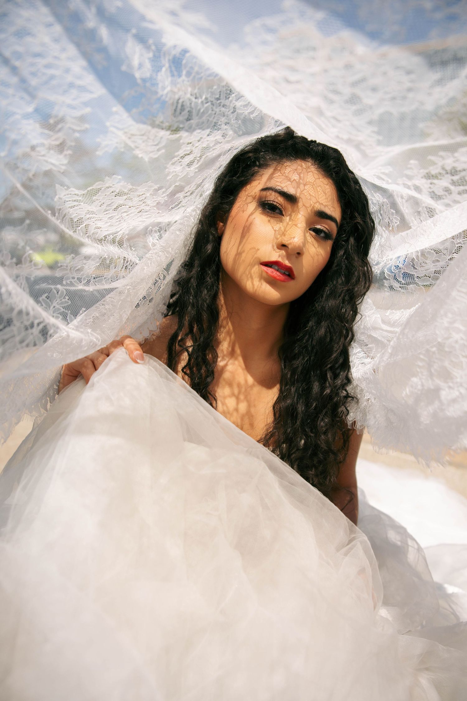 A bride with dark curly hair poses gracefully beneath a flowing white lace veil in soft dappled light.