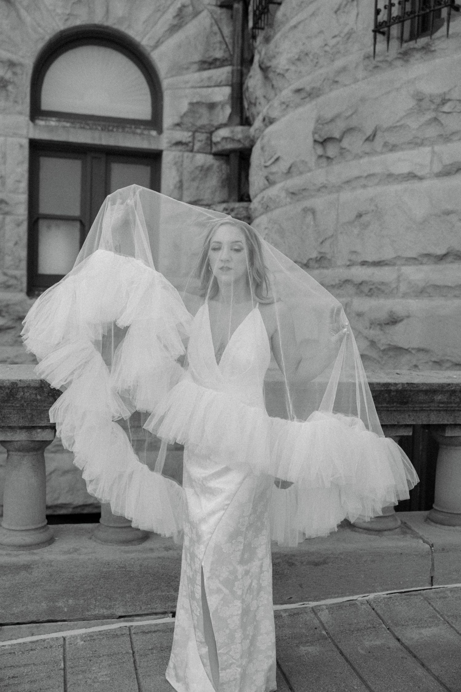 Bride in fitted lace gown with dramatic ruffled tulle veil flowing in wind before stone building, black and white.