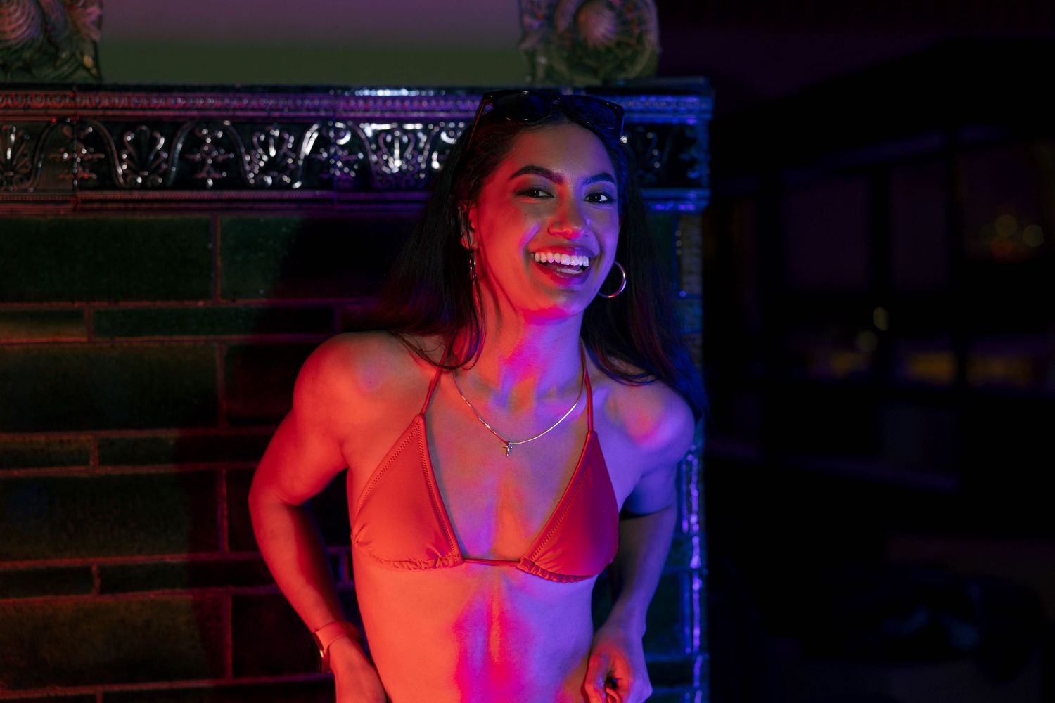 Smiling woman in a pink bikini top illuminated by colorful neon lights in a dark setting.