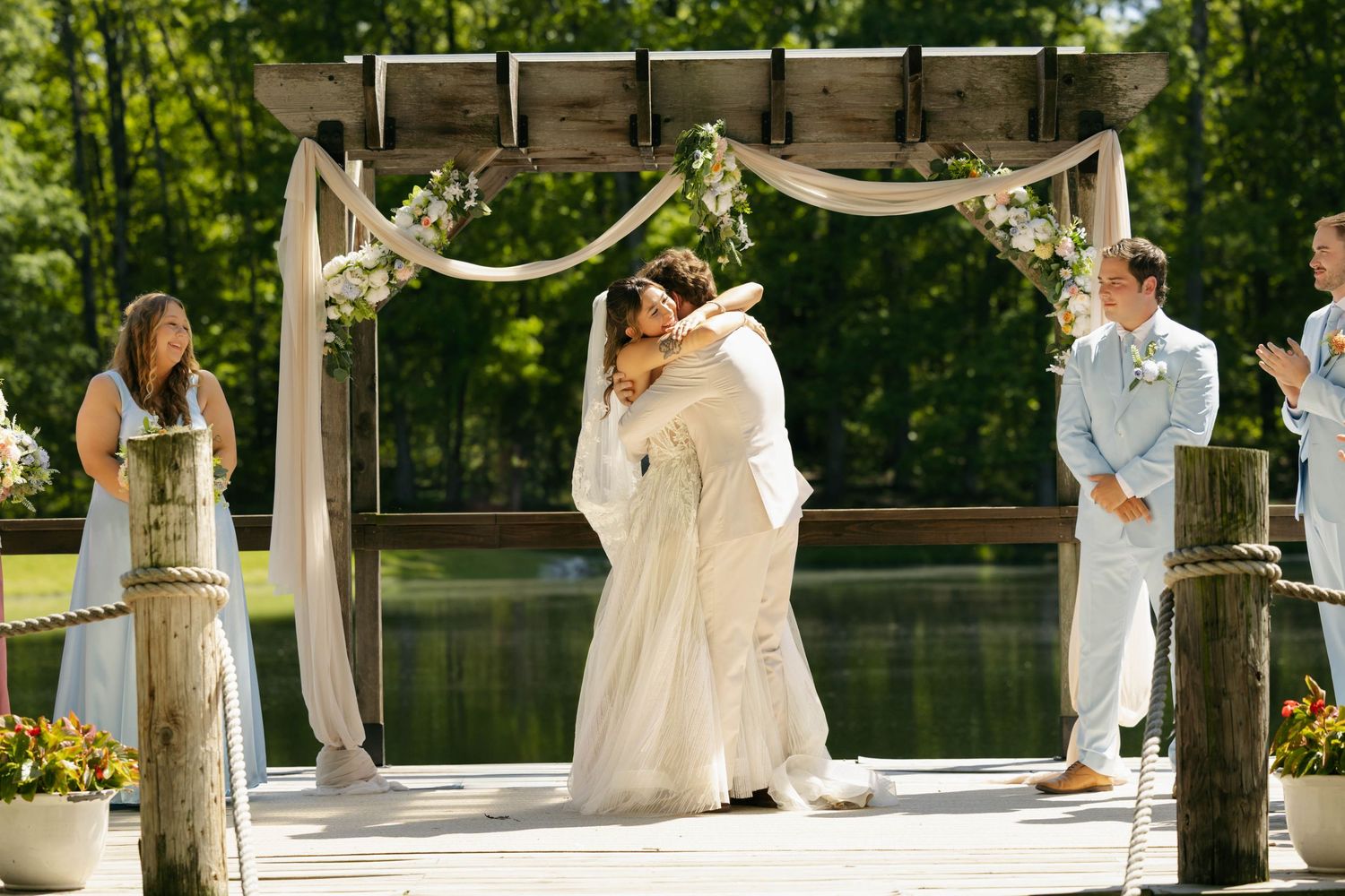 Bride and groom share first kiss under floral arch on dock at outdoor waterfront wedding ceremony.
