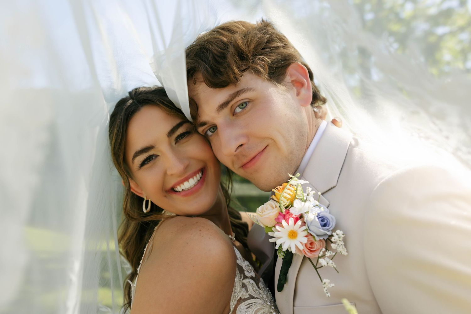 Smiling bride and groom posing under a white veil, groom wearing a floral boutonniere on his suit jacket.