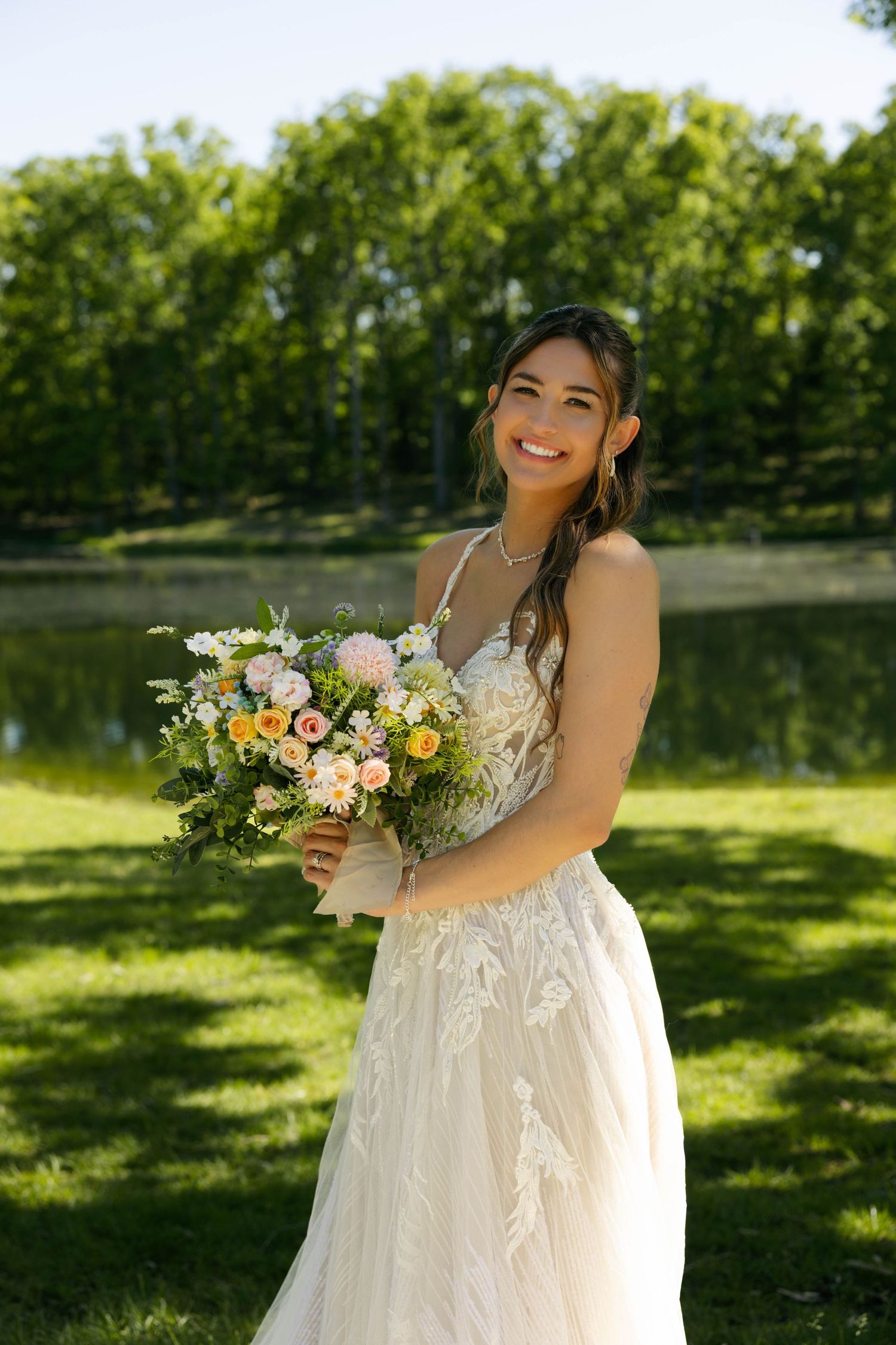 Smiling bride in lace gown holds colorful bouquet outdoors near pond with green trees.