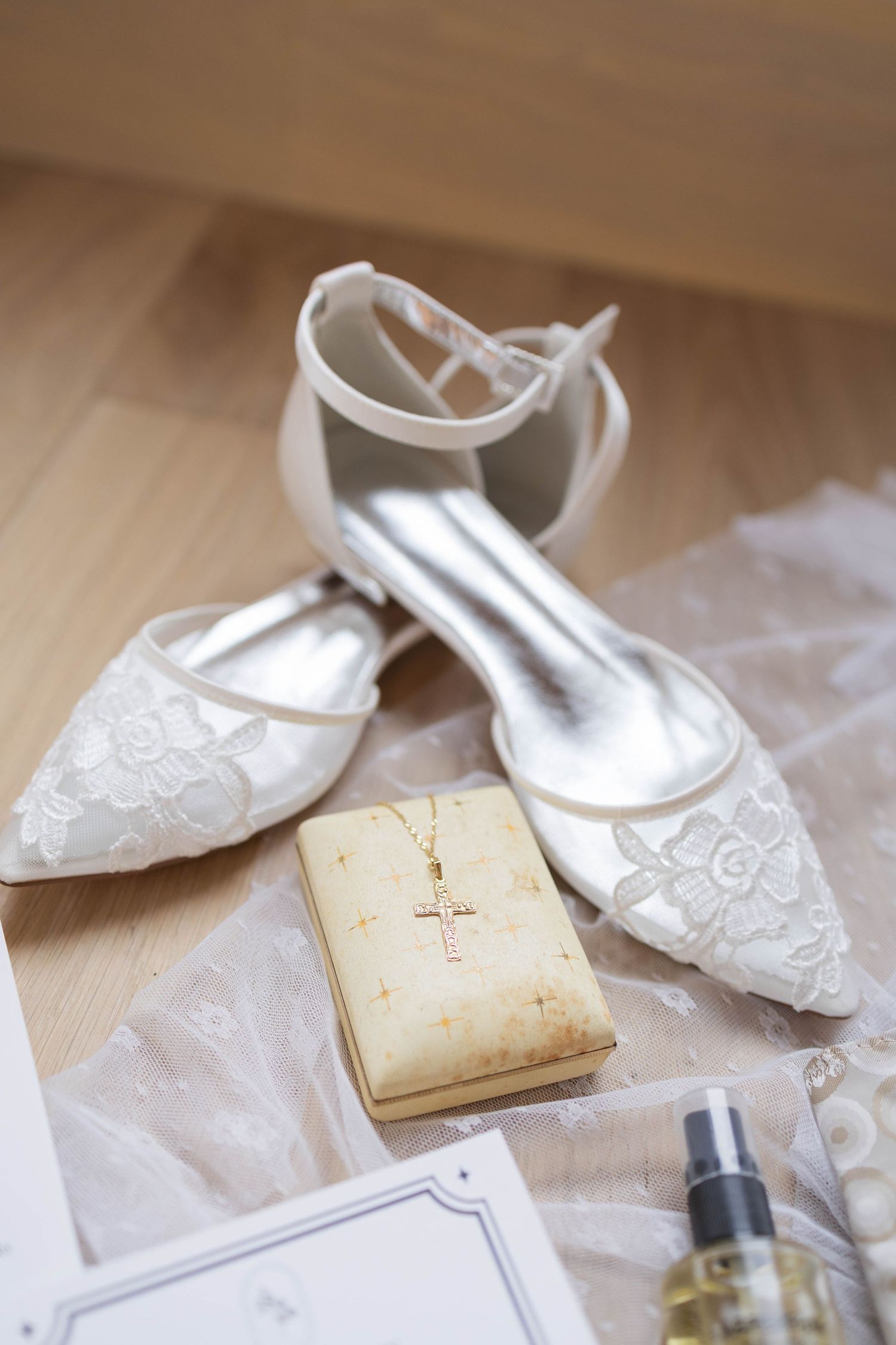 White lace bridal flats with ankle straps beside a gold cross necklace on a small book and perfume bottle.