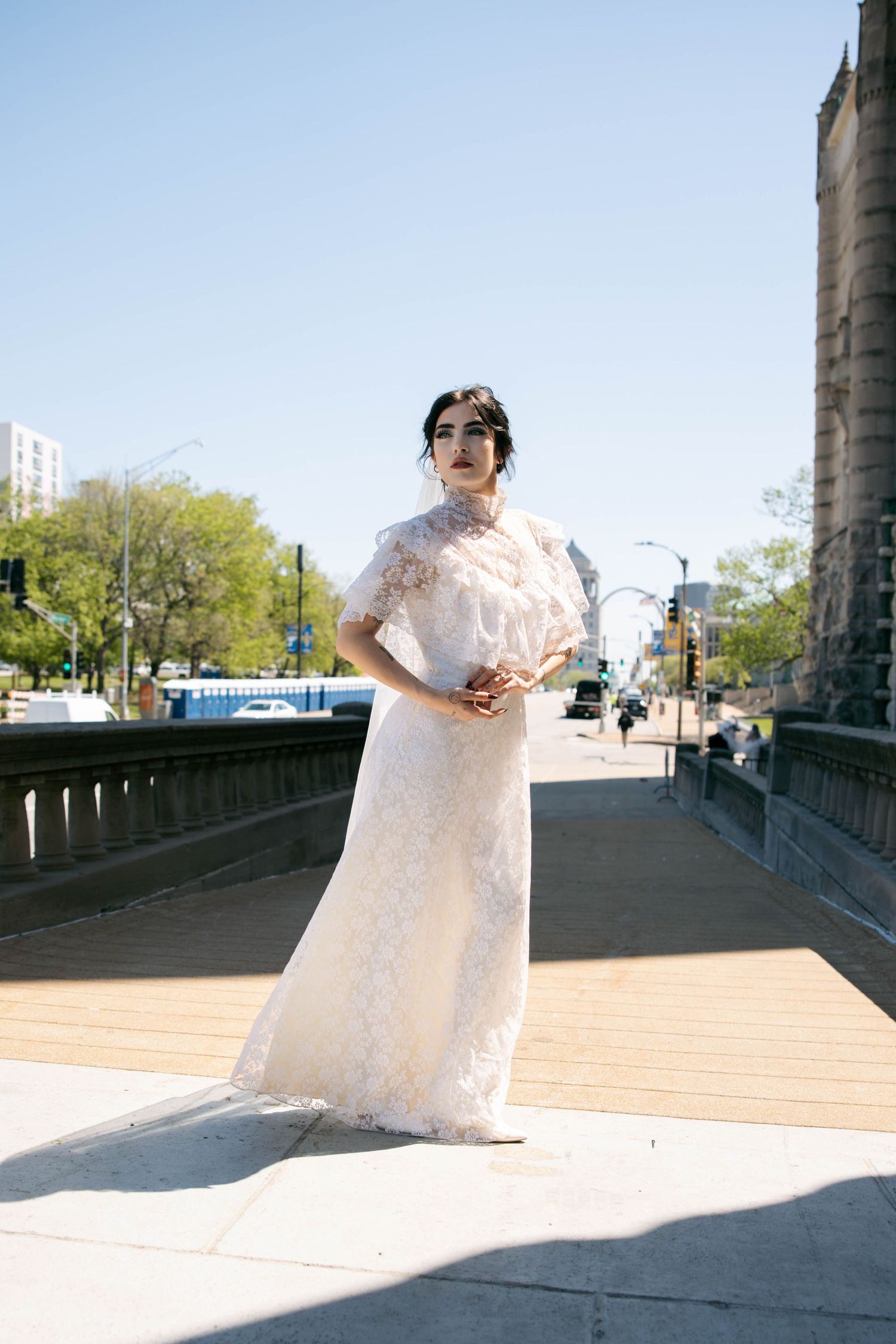 Bride in white lace gown poses on urban bridge on sunny day.