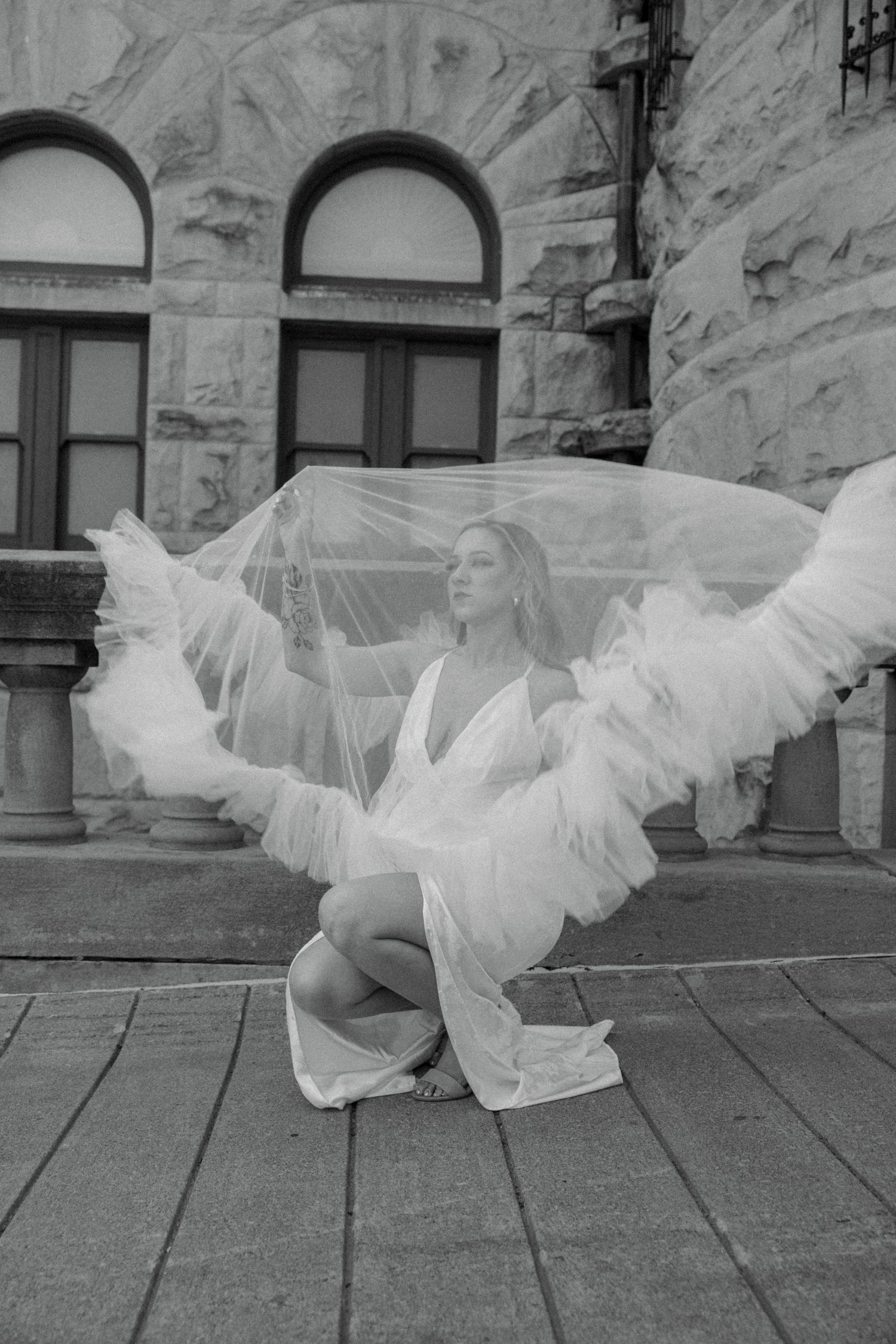 Bride in lace wedding gown sits on steps holding dramatic ruffled tulle veil aloft outside historic stone building, black and white.