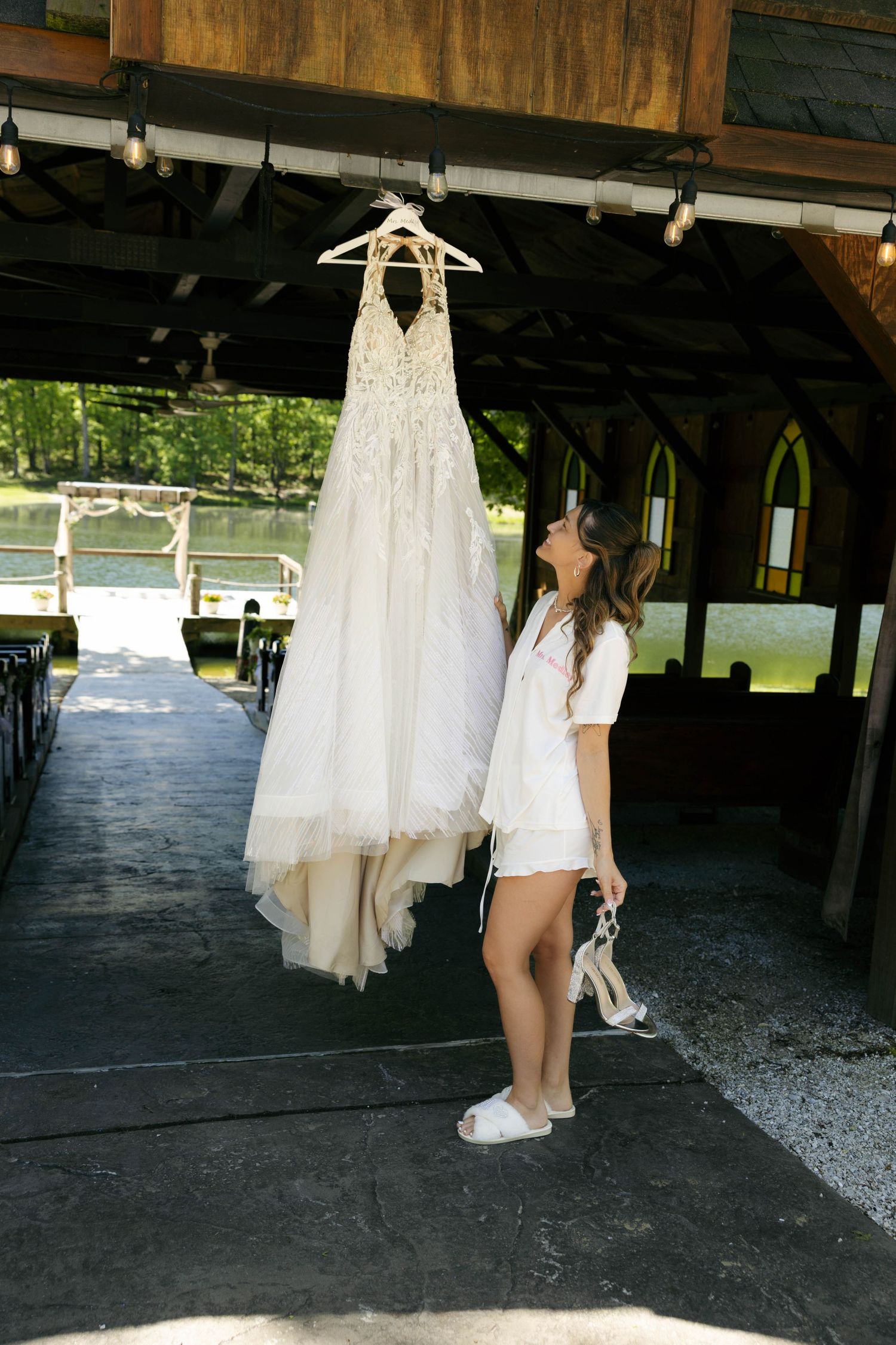 Bride in white robe admiring her lace wedding gown hanging outdoors at a rustic waterfront venue.