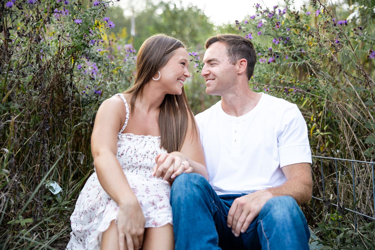 A romantic couple sits together among flowering bushes and greenery in casual outdoor portraits.