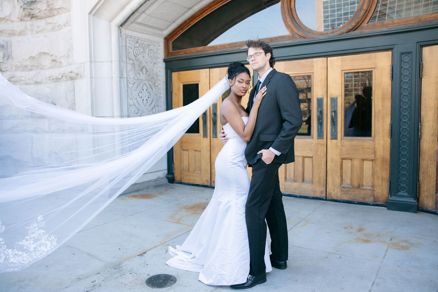 Couple embraces outside ornate church doors as bride's cathedral veil billows dramatically in the wind.