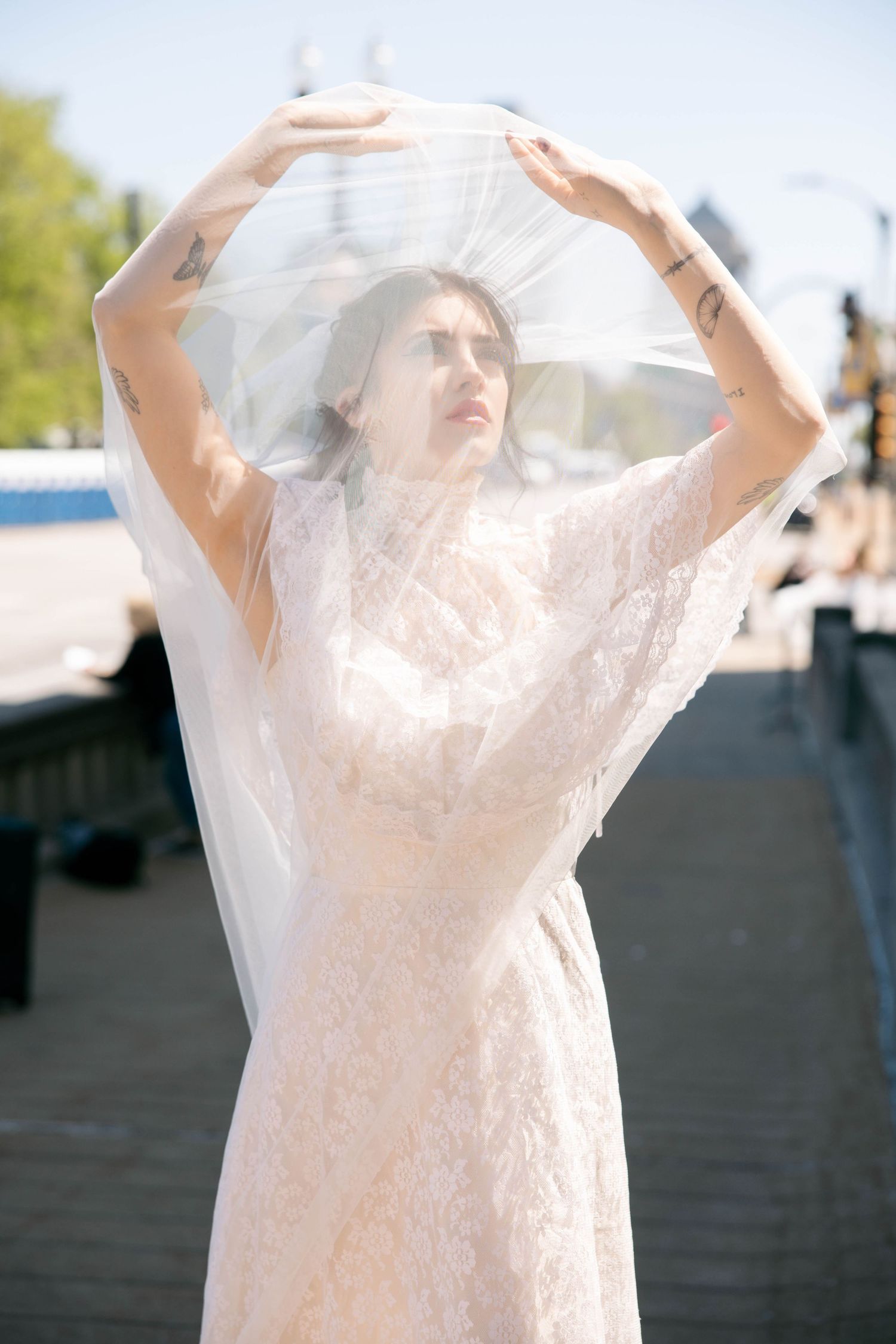 Tattooed bride in lace dress lifts sheer veil overhead on sunny urban street.
