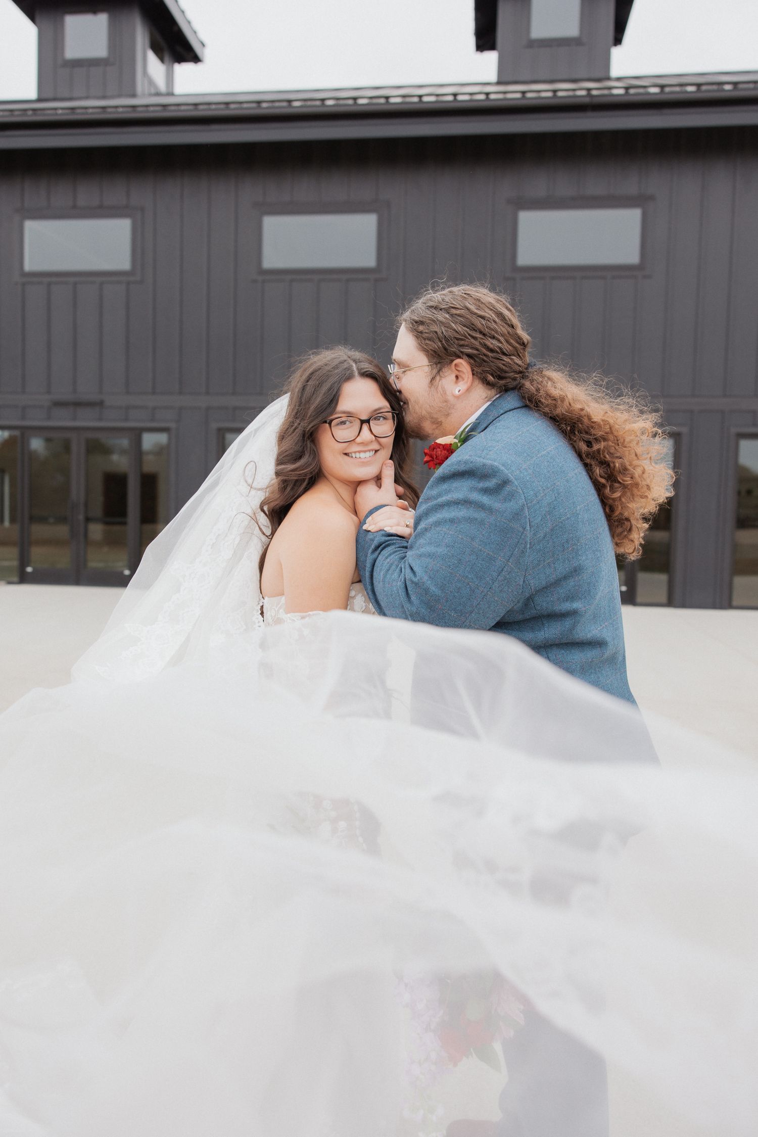 Couple sharing an intimate moment under a flowing white veil against a modern gray building.