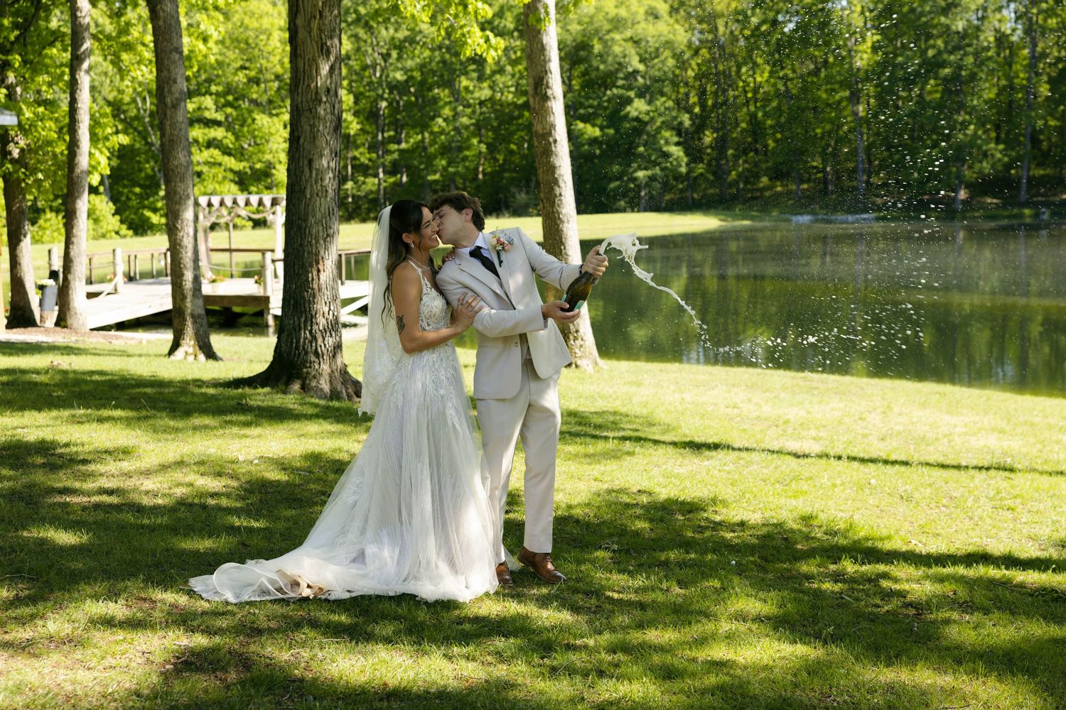 Bride and groom kissing while popping champagne bottle outdoors near a pond surrounded by lush green trees.