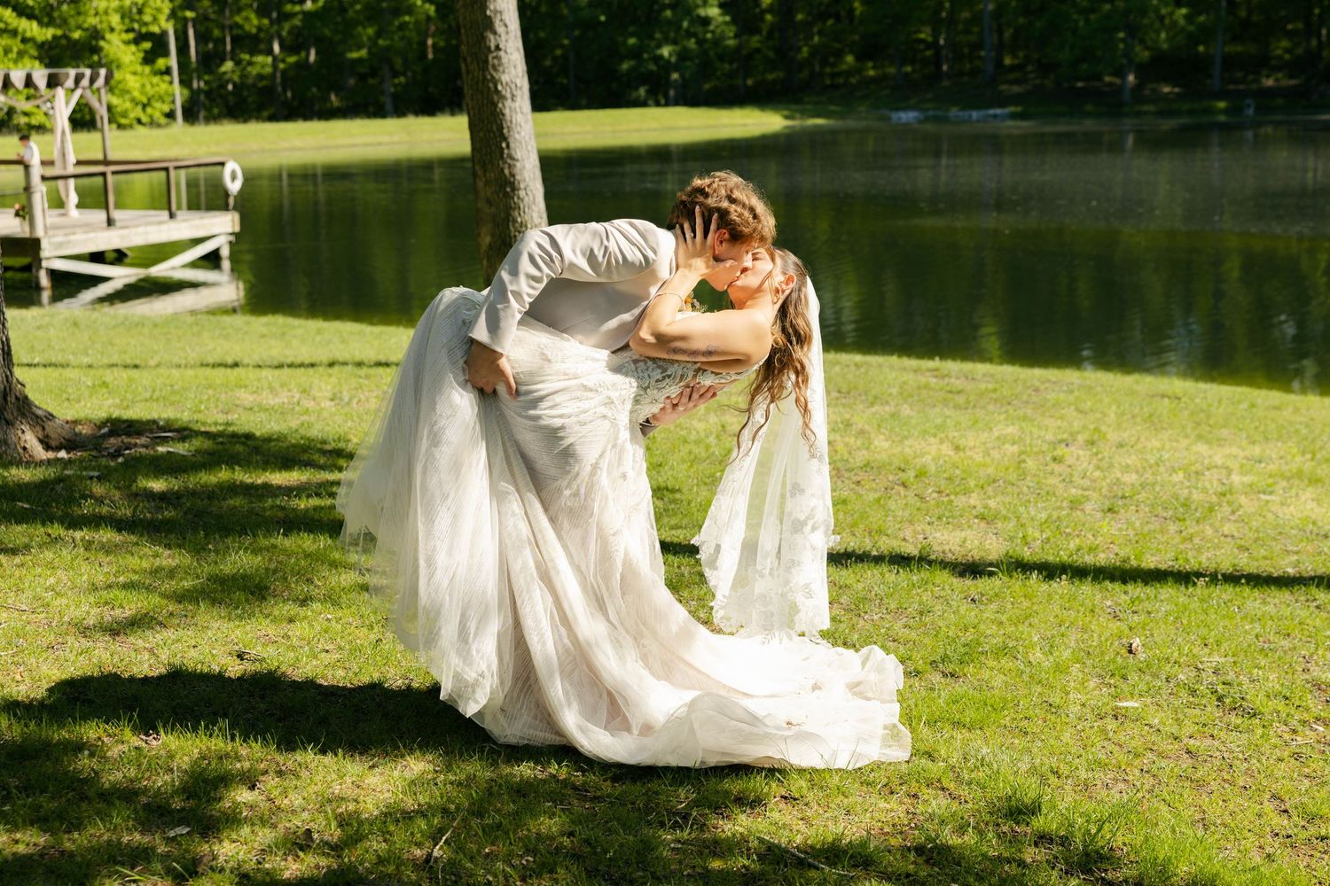 A bride and groom share a romantic dip kiss in white wedding attire on a lush green lawn beside a serene lake.