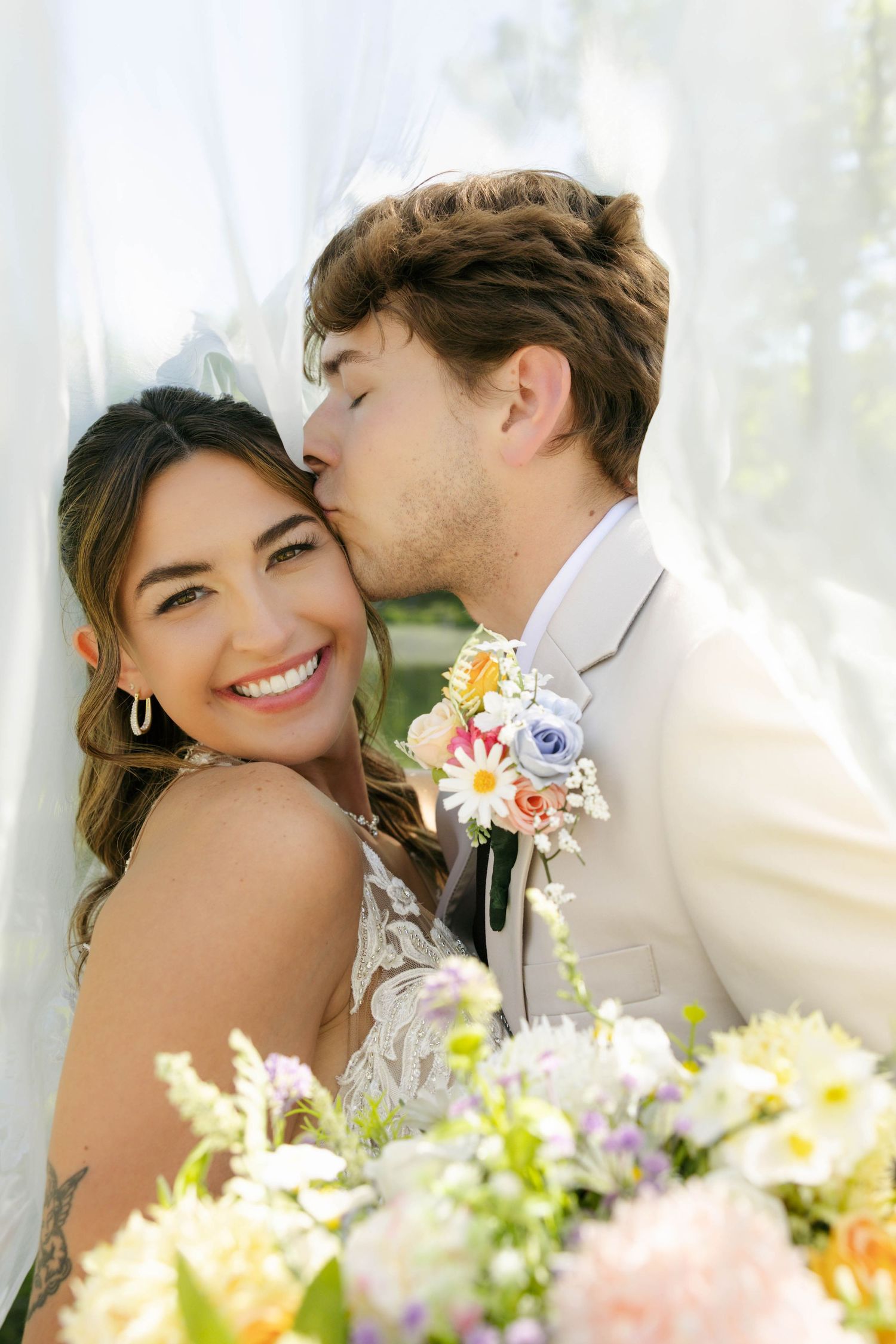 Groom in white suit kissing smiling bride on cheek under veil with colorful floral bouquet in foreground.