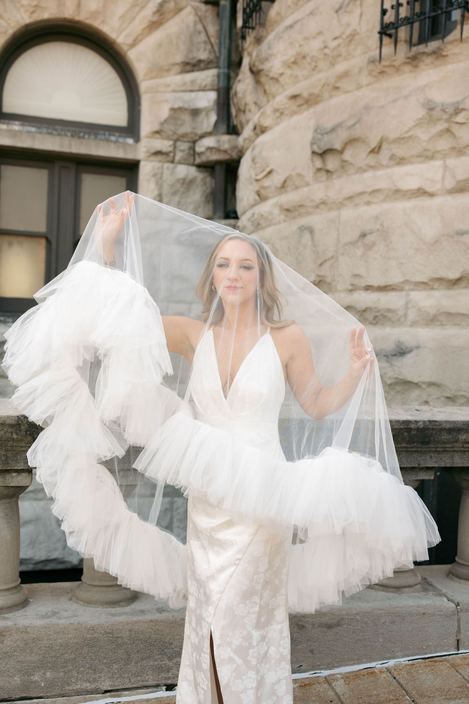 Elegant bride in sleek lace wedding dress holds billowing ruffled tulle veil open wide outside historic stone building.