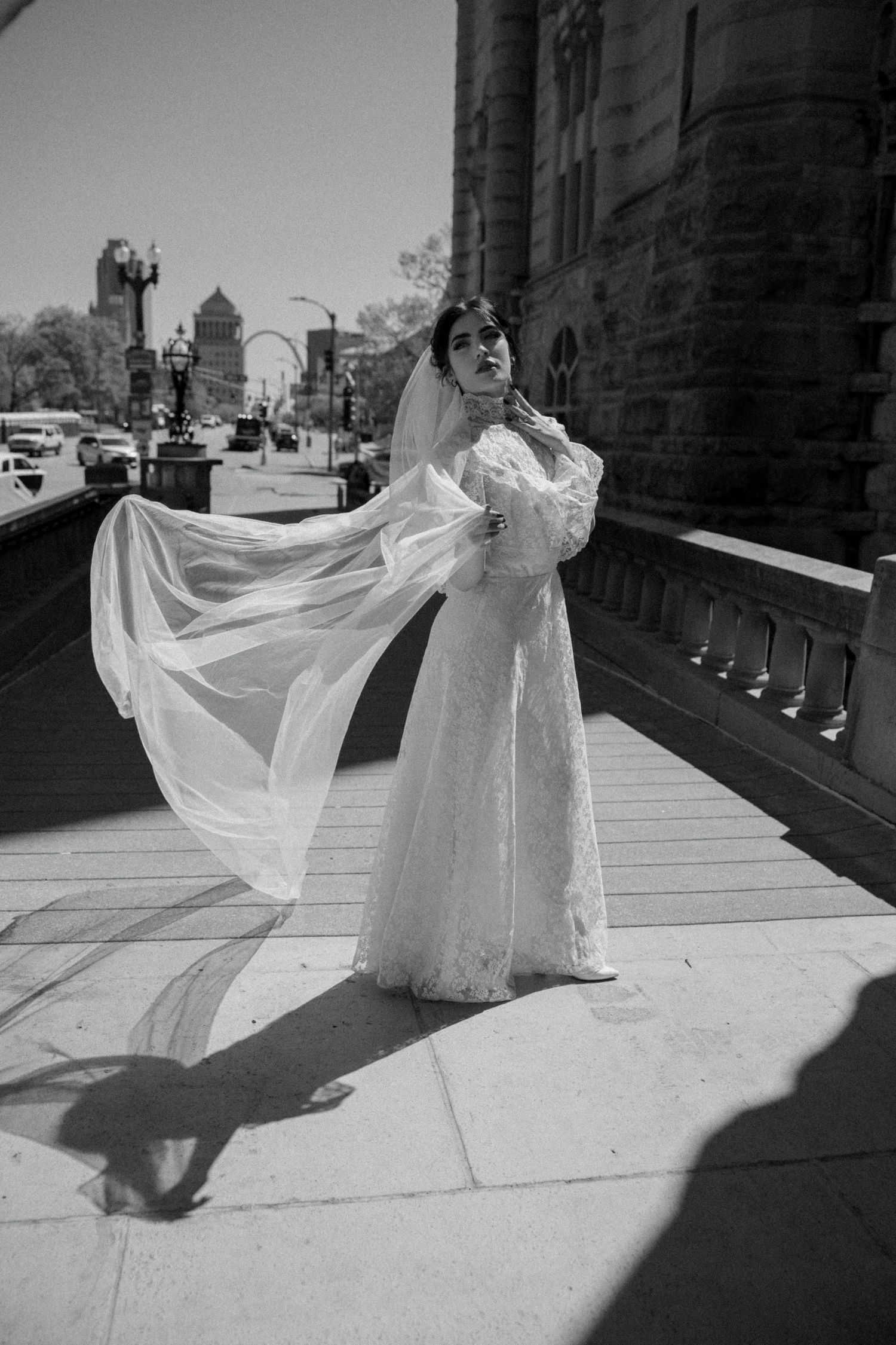 Black and white photo of a bride in lace gown with flowing veil on urban balcony.