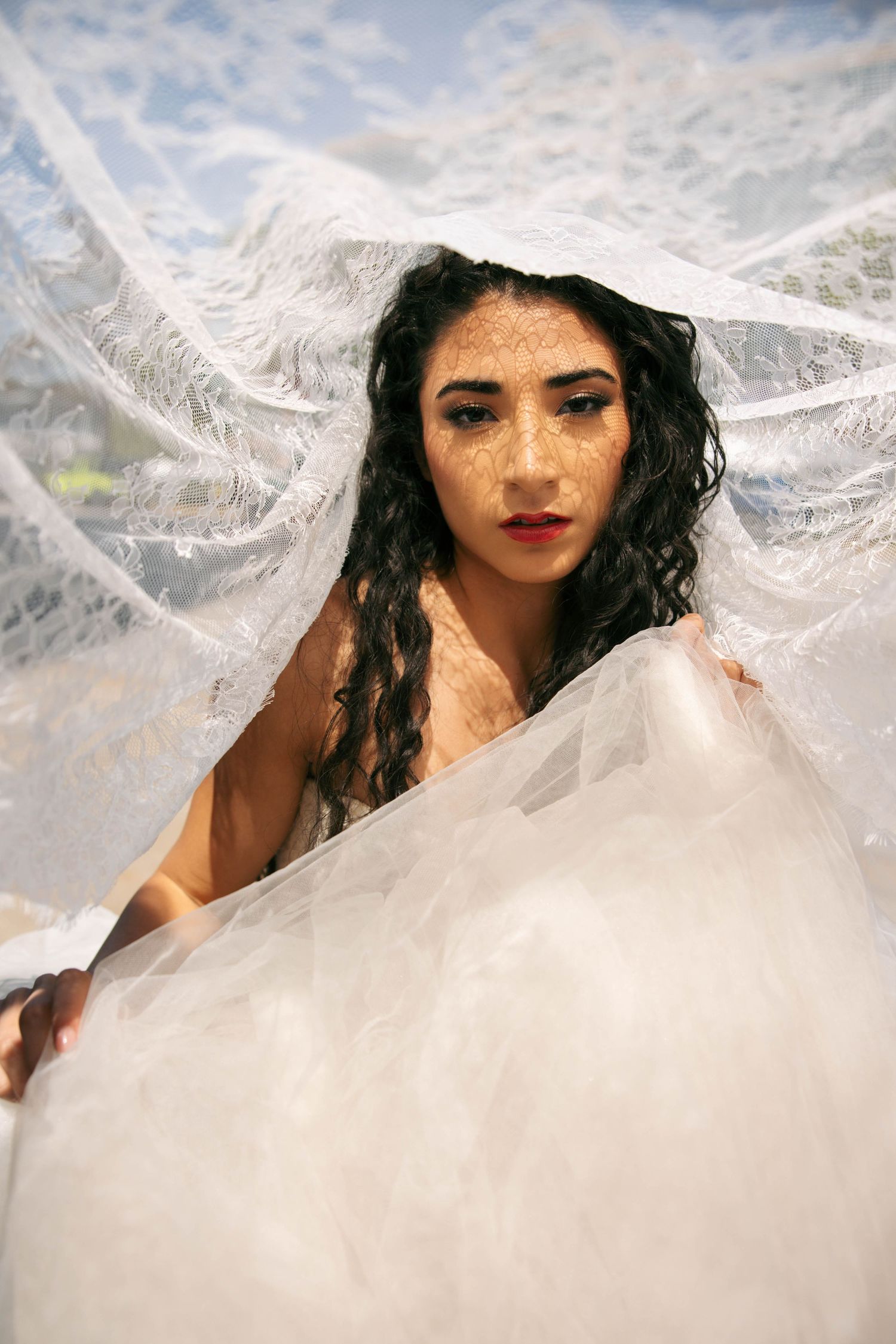 Woman in white wedding dress holding sheer lace veil overhead, gazing forward in outdoor setting.