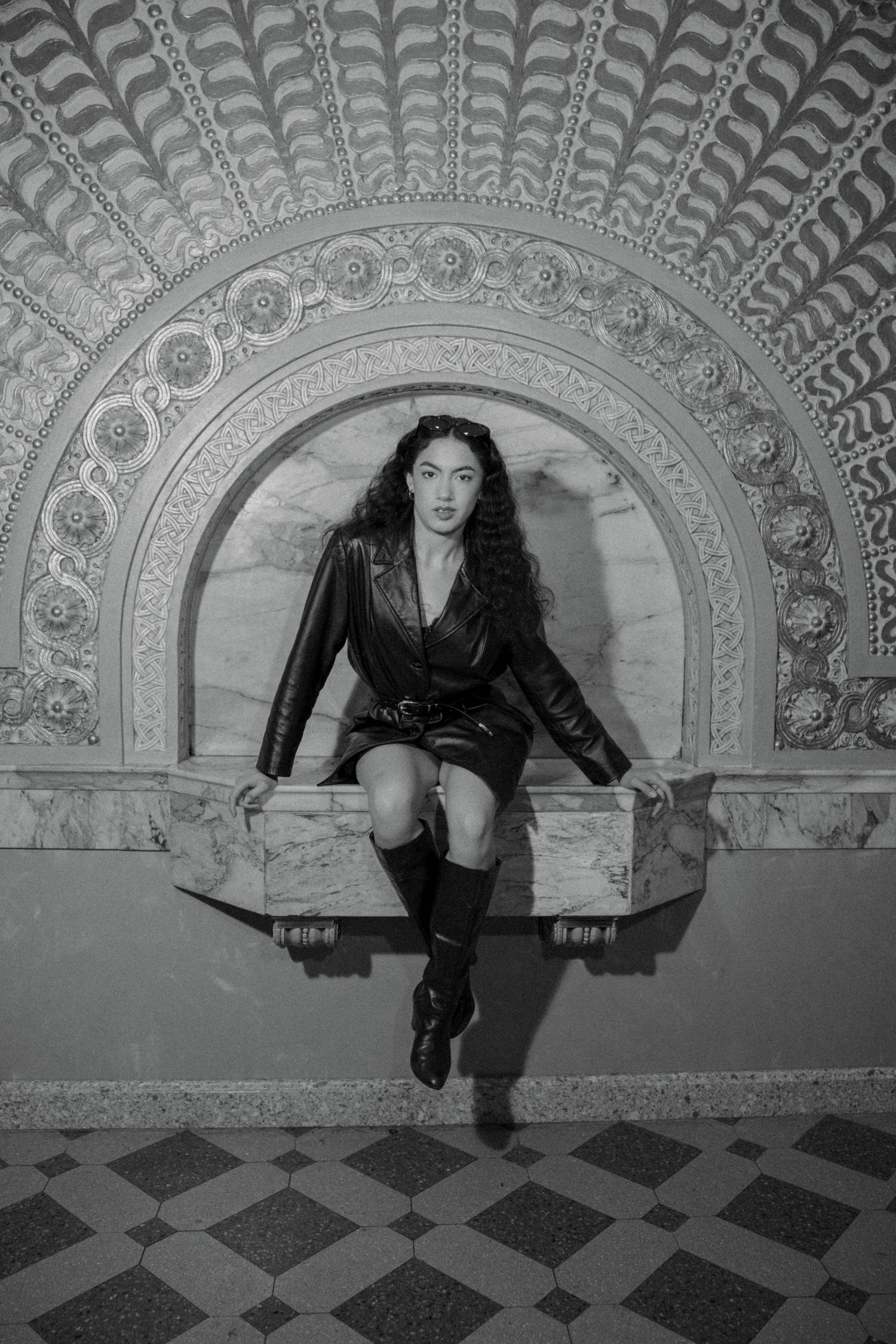 Black and white photo of a woman in leather dress and boots sitting in ornate architectural niche.