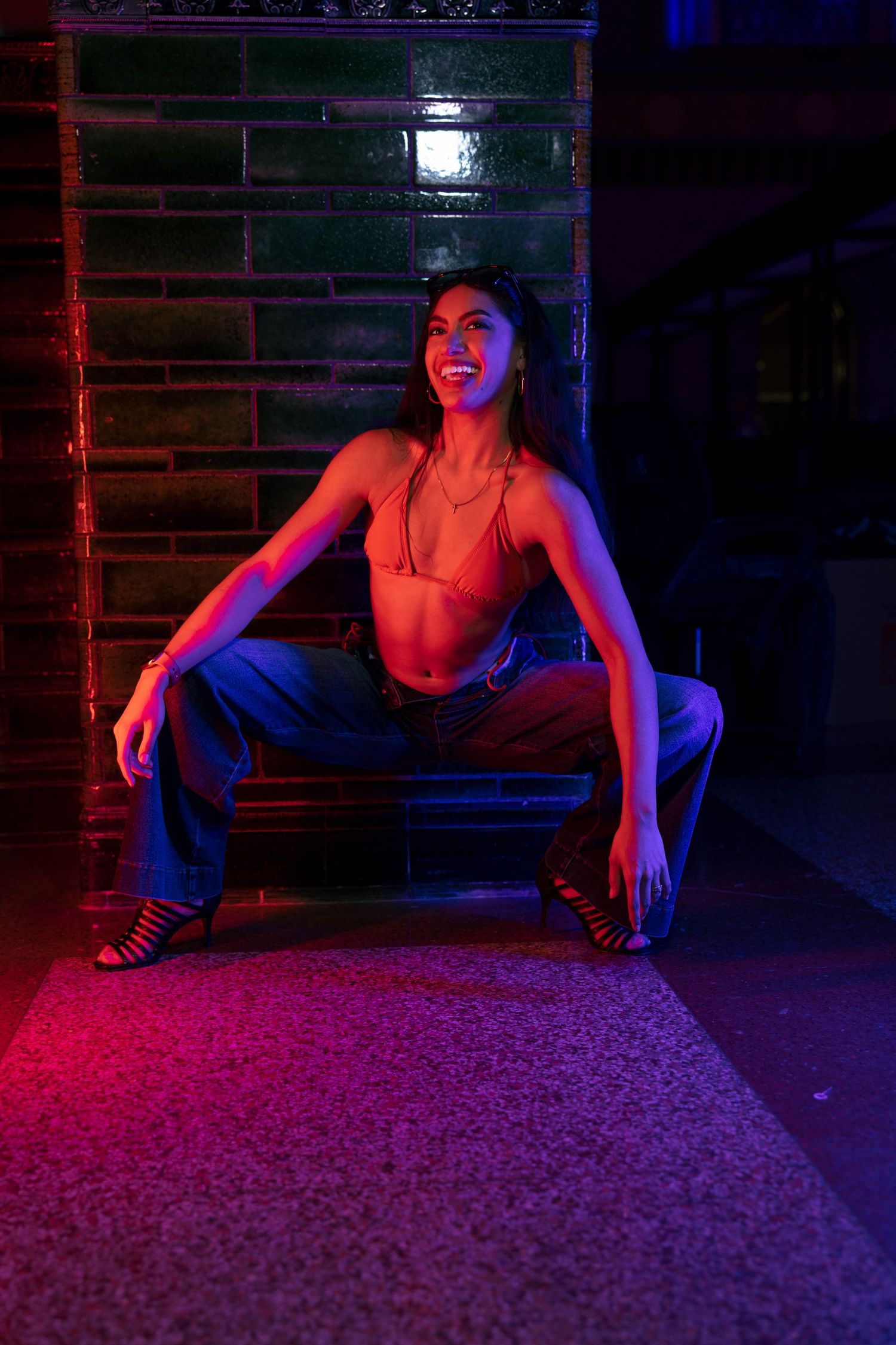 Woman in bikini top and jeans posing under neon lights against a brick wall at night.