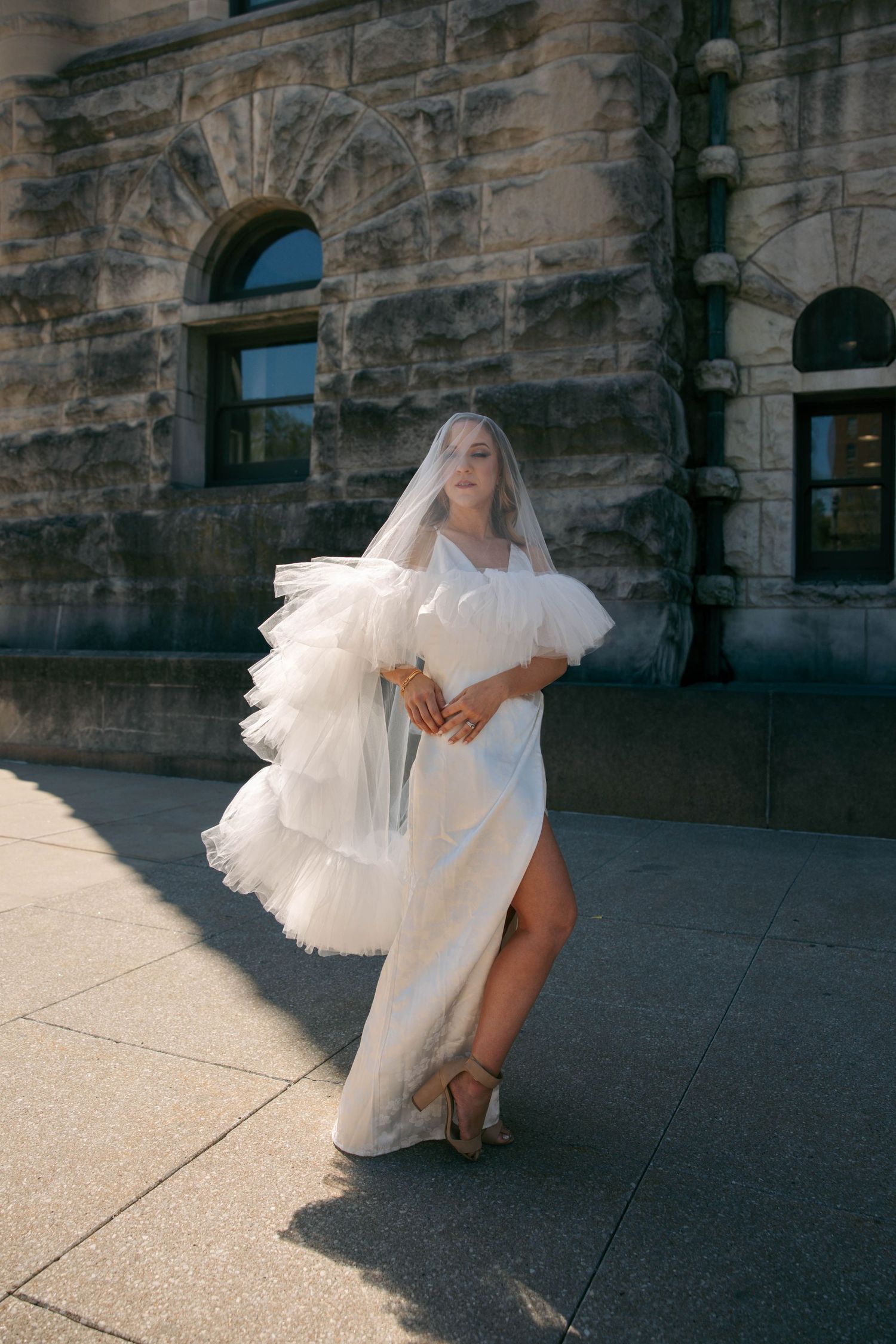 Bride in fitted white gown with ruffled tulle veil walks in sunlight outside grand stone building holding bouquet.