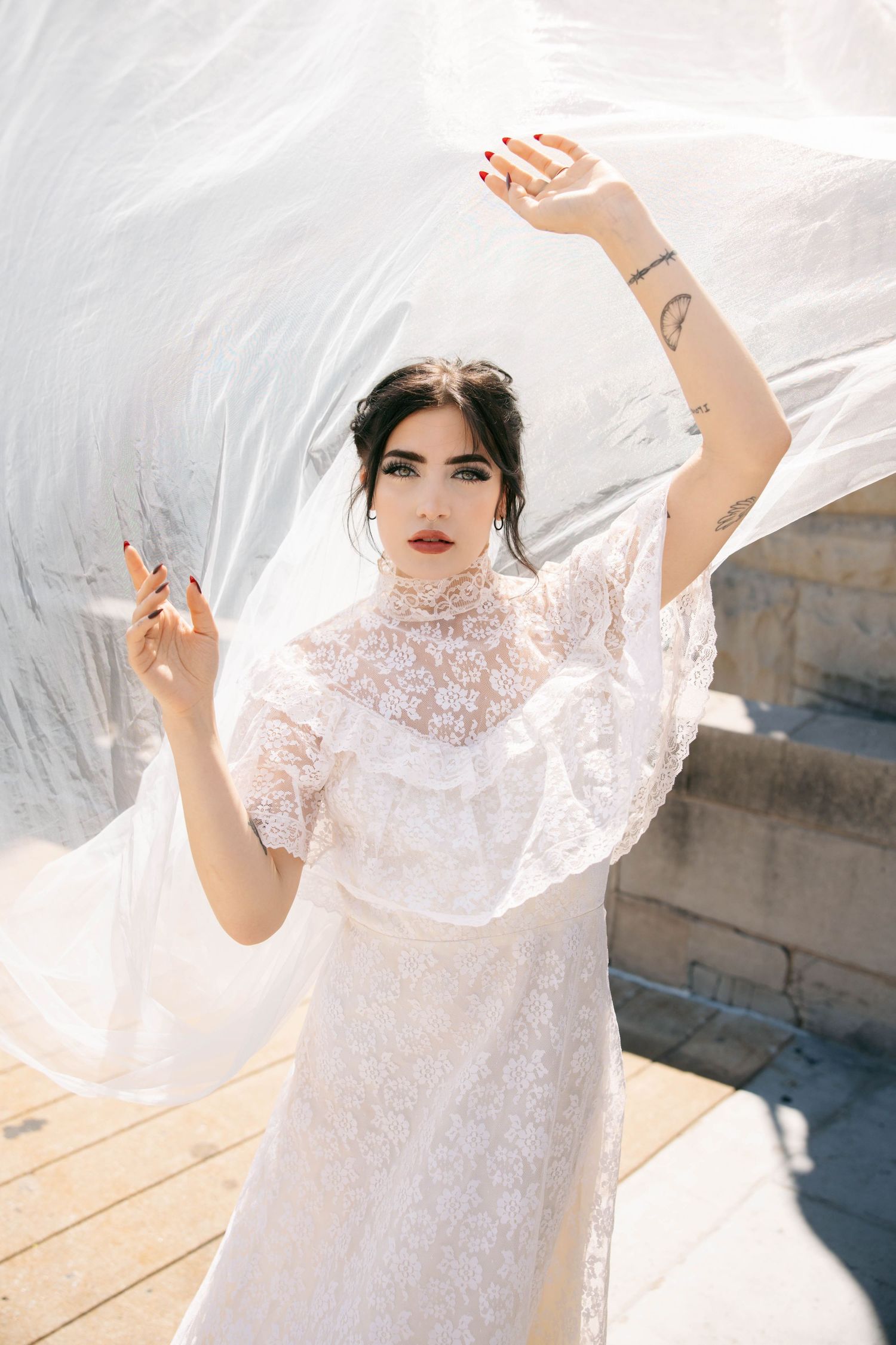 Bride in white lace dress lifting veil with tattooed arms in bright outdoor setting.