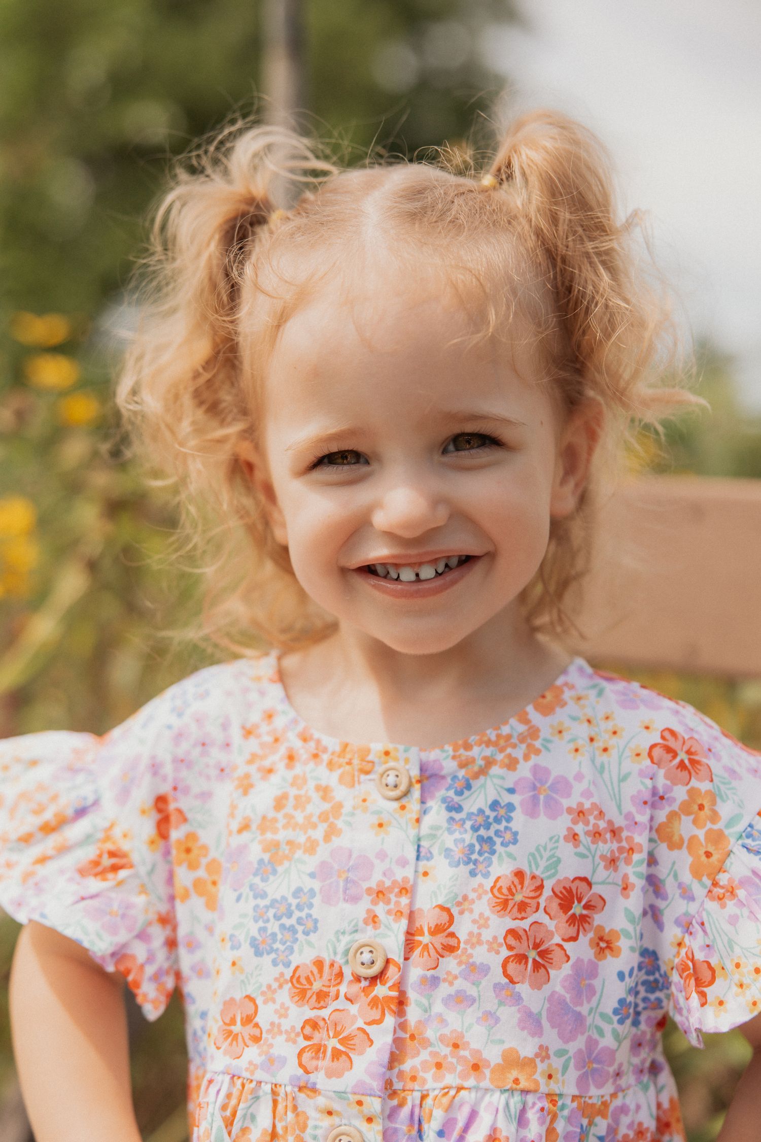 Child in floral print dress smiles brightly in outdoor garden setting.