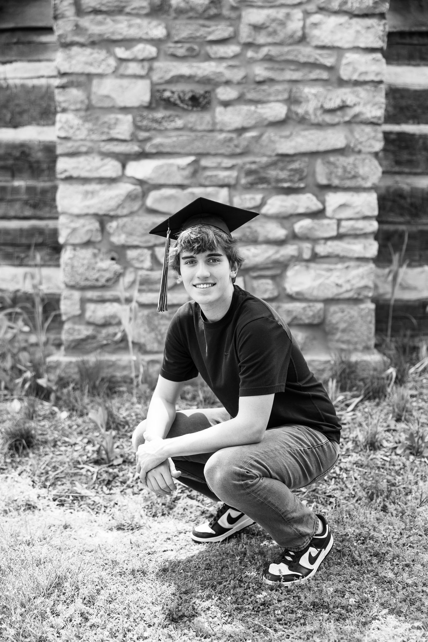A graduate in cap and black t-shirt crouches by a stone wall in black and white photography.
