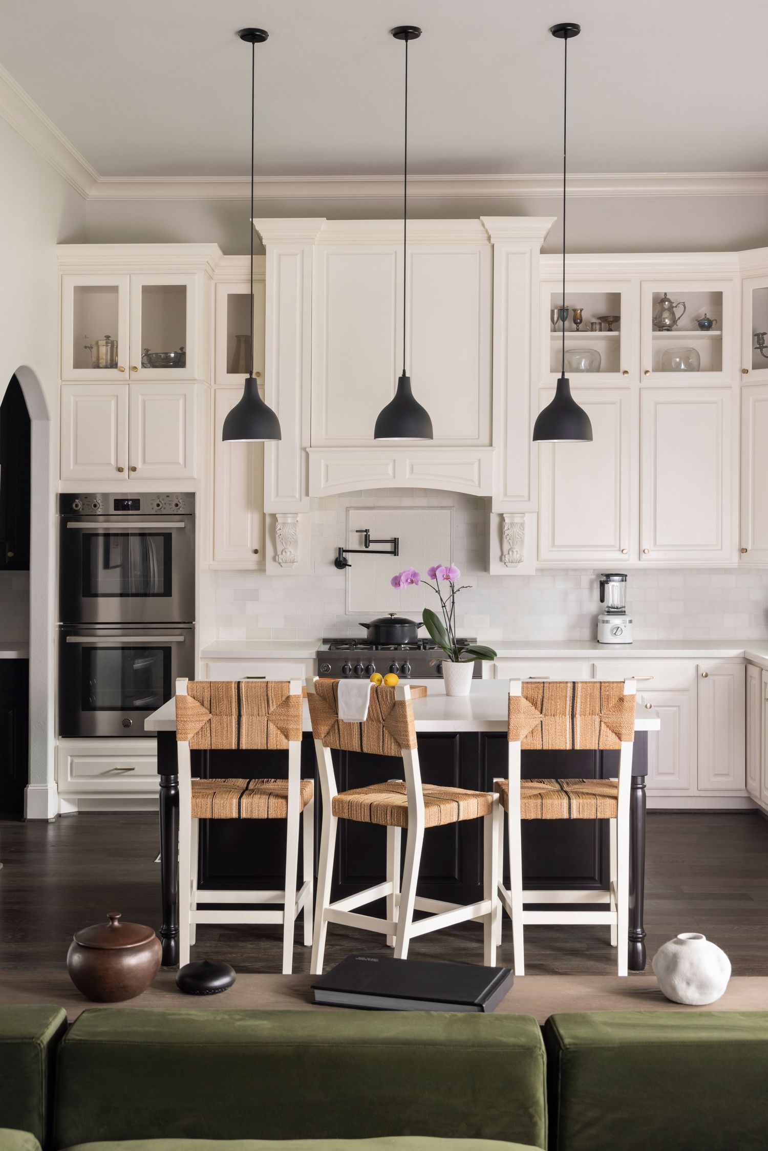 Modern white kitchen with olive green sectional sofa, wood barstools, and pendant lighting over island.