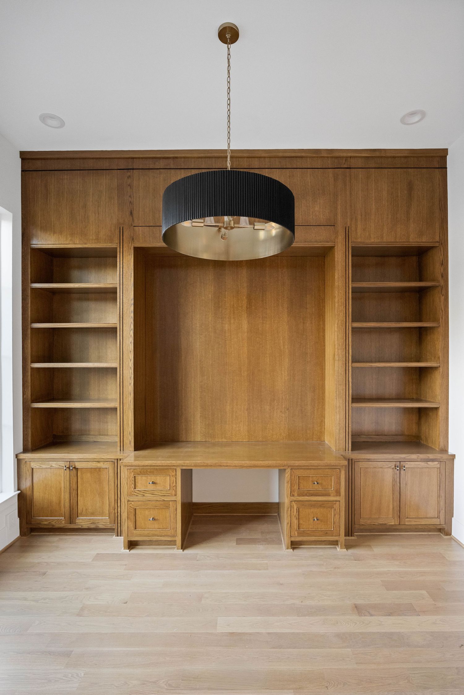 Built-in wooden office desk with shelving units and pendant lighting on light hardwood floors.