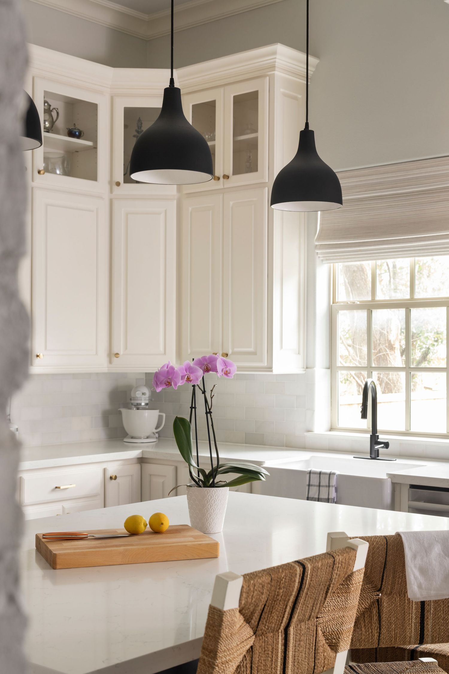 Modern white kitchen with farmhouse sink, pendant lights, and woven rattan barstools at a marble counter.